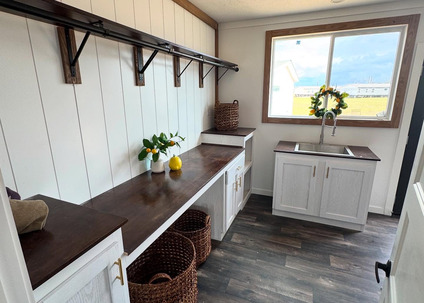 Bright laundry room with large window, dark wood countertops, white cabinets, and wicker baskets. Fresh plants and a lemon add a touch of nature.