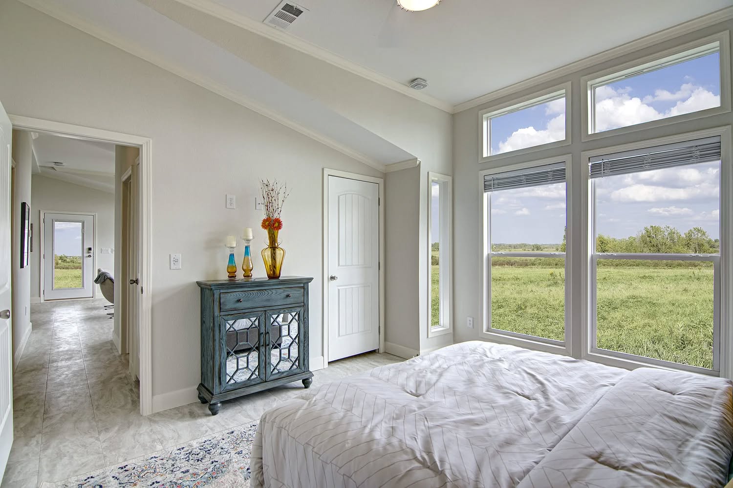 A bright bedroom with large windows showing a grassy landscape. Features a bed, a blue decorative cabinet with vases, and a neutral, calm tone.