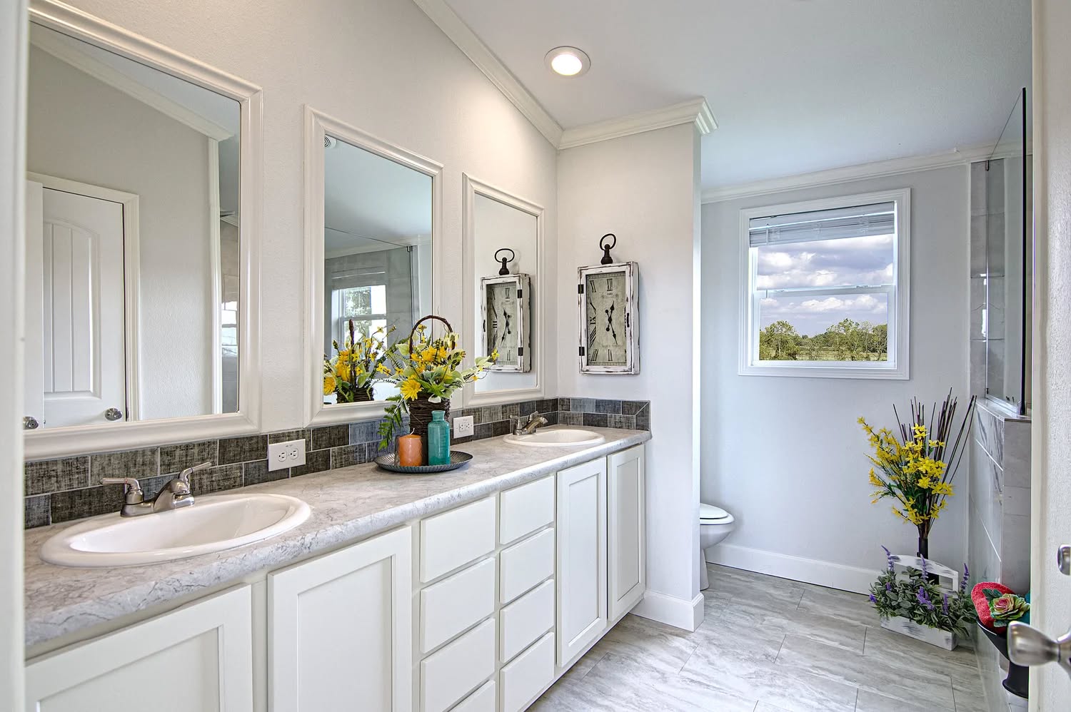 Bright bathroom with two sinks, large mirrors, and a window showing a view of trees. Yellow flowers provide a fresh, inviting touch.