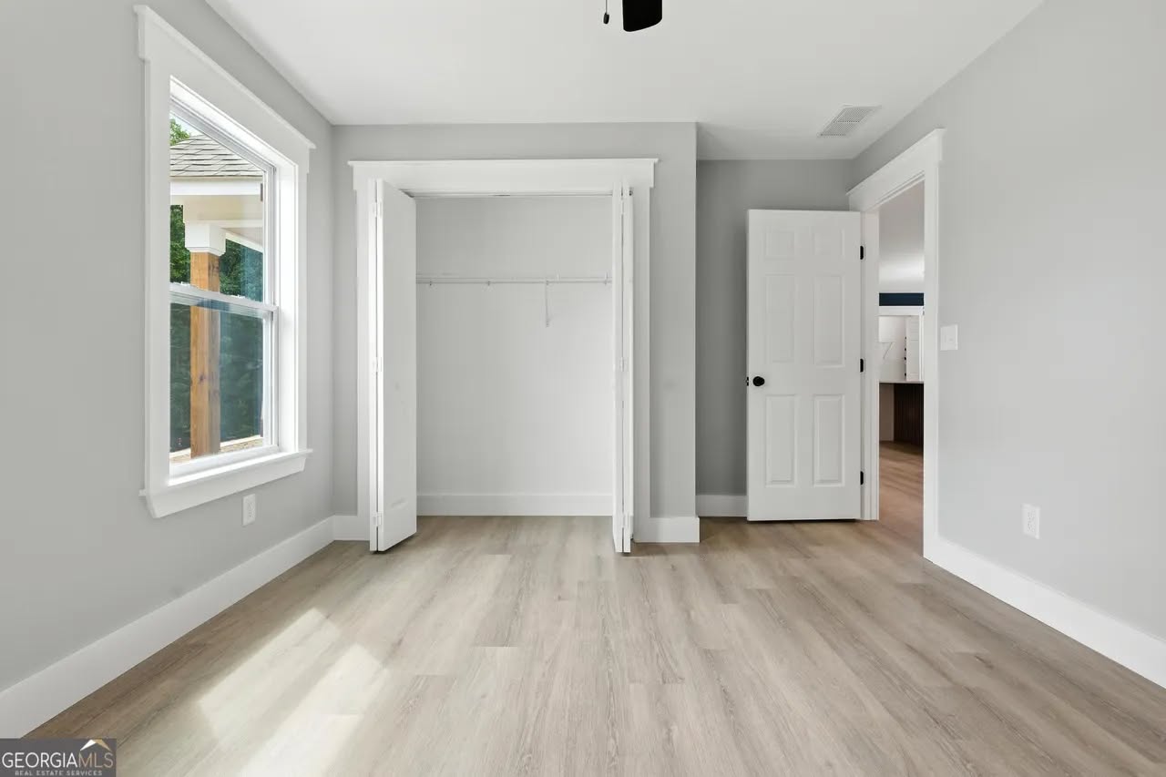 Bright, empty room with light wood flooring, gray walls, and a large window. Open closet with white doors and an adjacent open door showing another room.