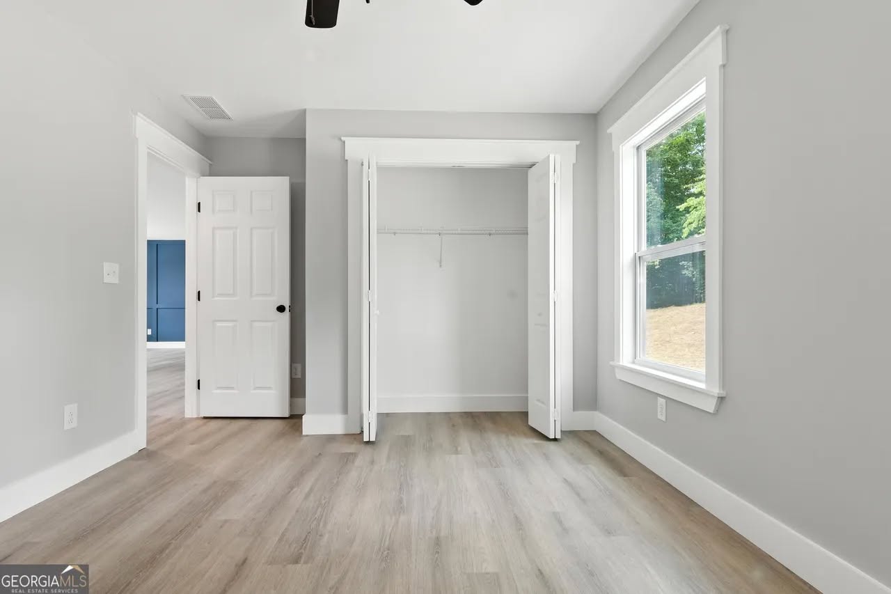 Bright, empty room with light gray walls and wooden floors, featuring an open closet with white doors and a large window letting in natural light.