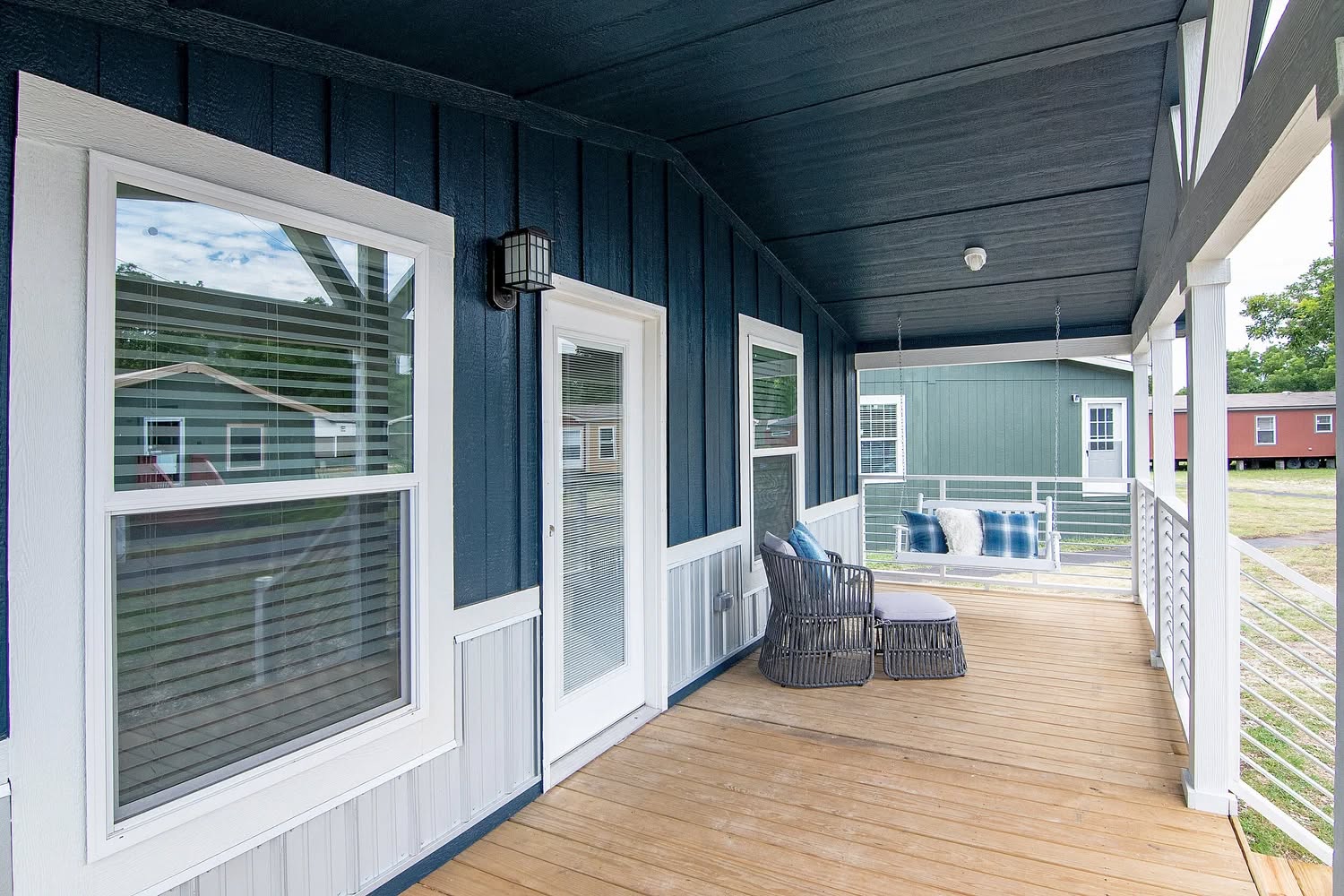 A cozy porch features a dark blue wooden wall with white-framed windows and a door. A cushioned chair and a swing with blue pillows add comfort.