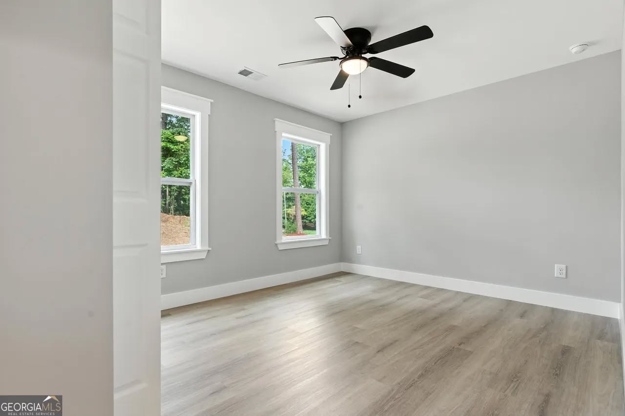 A bright, empty room with light wood flooring and gray walls. Two windows show a green outdoor scene. A black ceiling fan enhances the modern, airy feel.