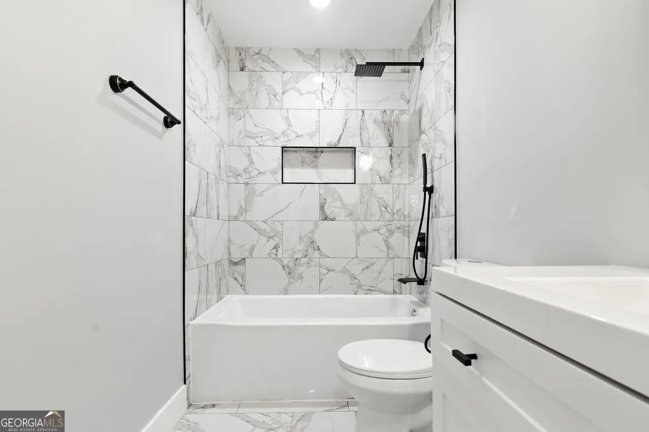 A modern bathroom featuring a white bathtub with black fixtures, set against marble-patterned tiles. The space is bright, minimalist, and sleek.