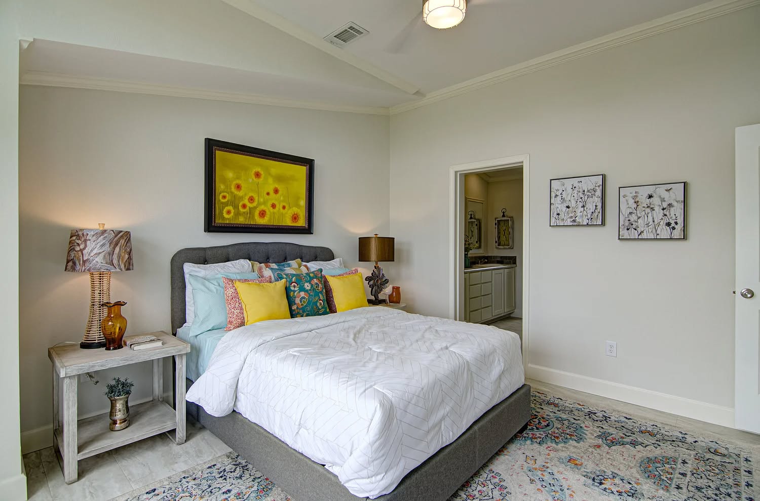 A cozy bedroom with a plush, gray bed adorned with colorful pillows and a white comforter. A bedside table holds a lamp and decor. Above the bed is a sunflower painting. The room has a soft, inviting ambiance.