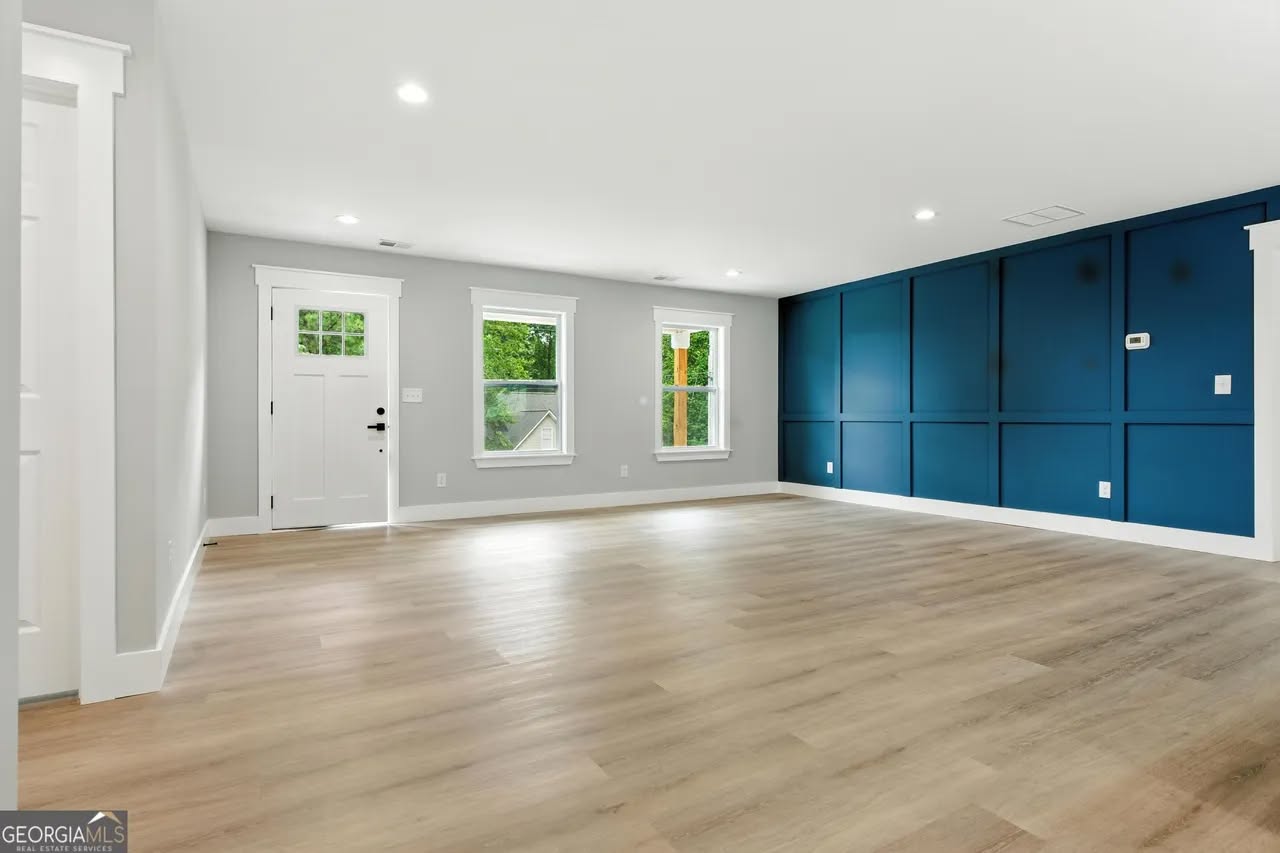 A spacious, empty room with light wooden floors, a bold blue accent wall, and three white-framed windows. A white door adds a crisp, modern touch.