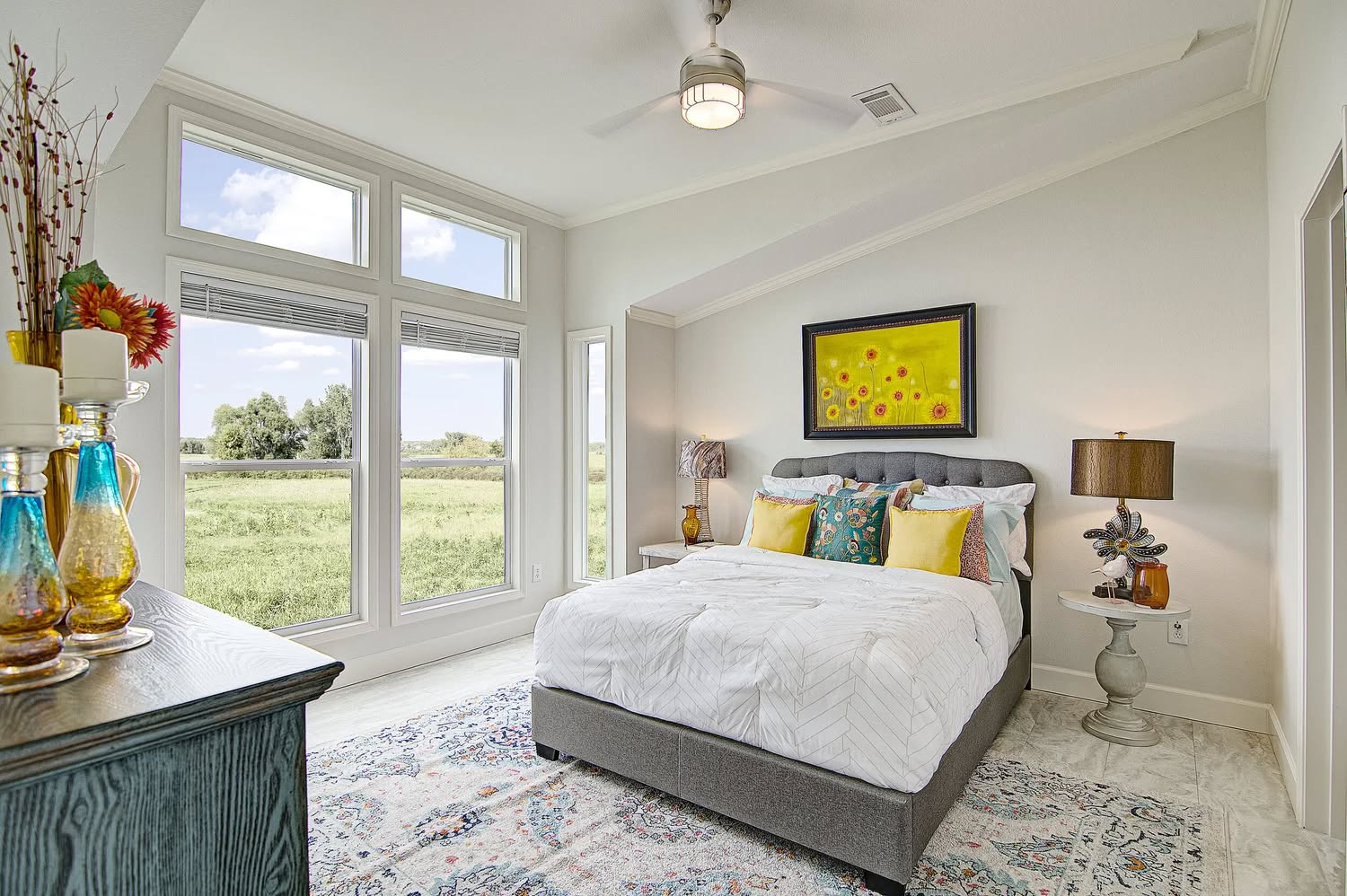A bright bedroom with a large window overlooking green fields. It features a gray bed, colorful pillows, floral painting, and vibrant decor on the dresser.