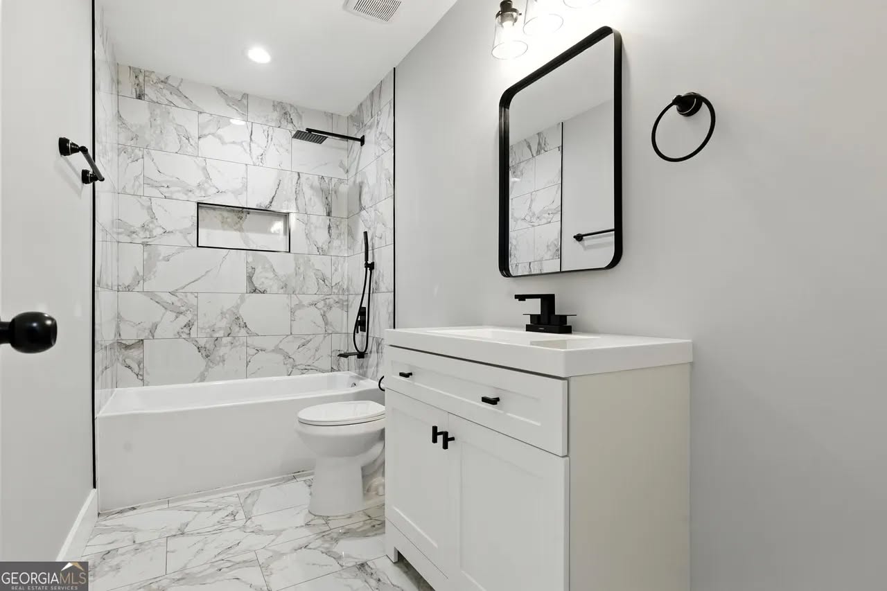 Modern bathroom with marble-tiled walls and floor, featuring a bathtub, toilet, and white vanity with a black-framed mirror. Bright, minimalist design.