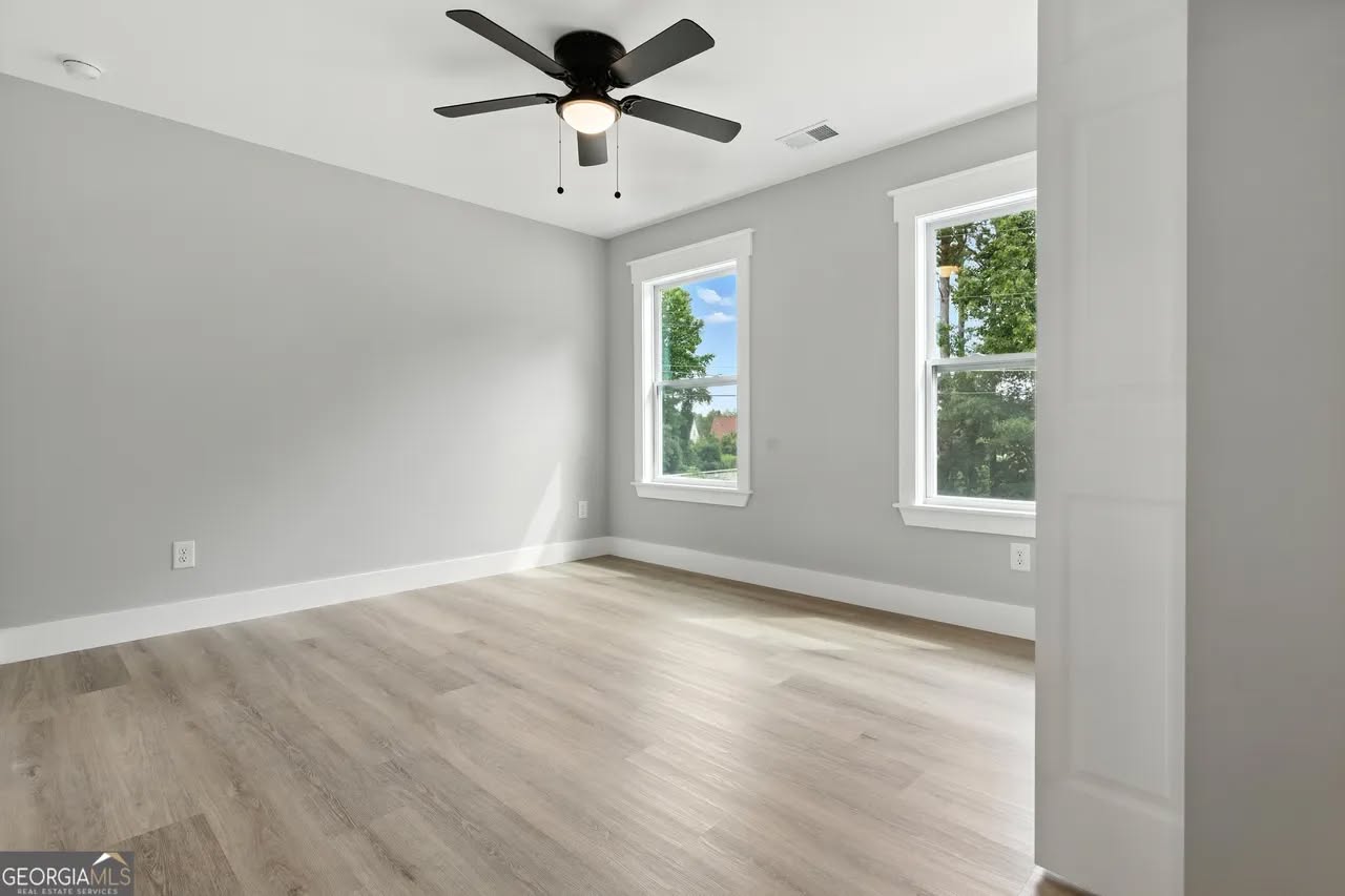 Empty room with light gray walls and hardwood floors, featuring two windows showing green trees. A black ceiling fan is centered on the white ceiling.