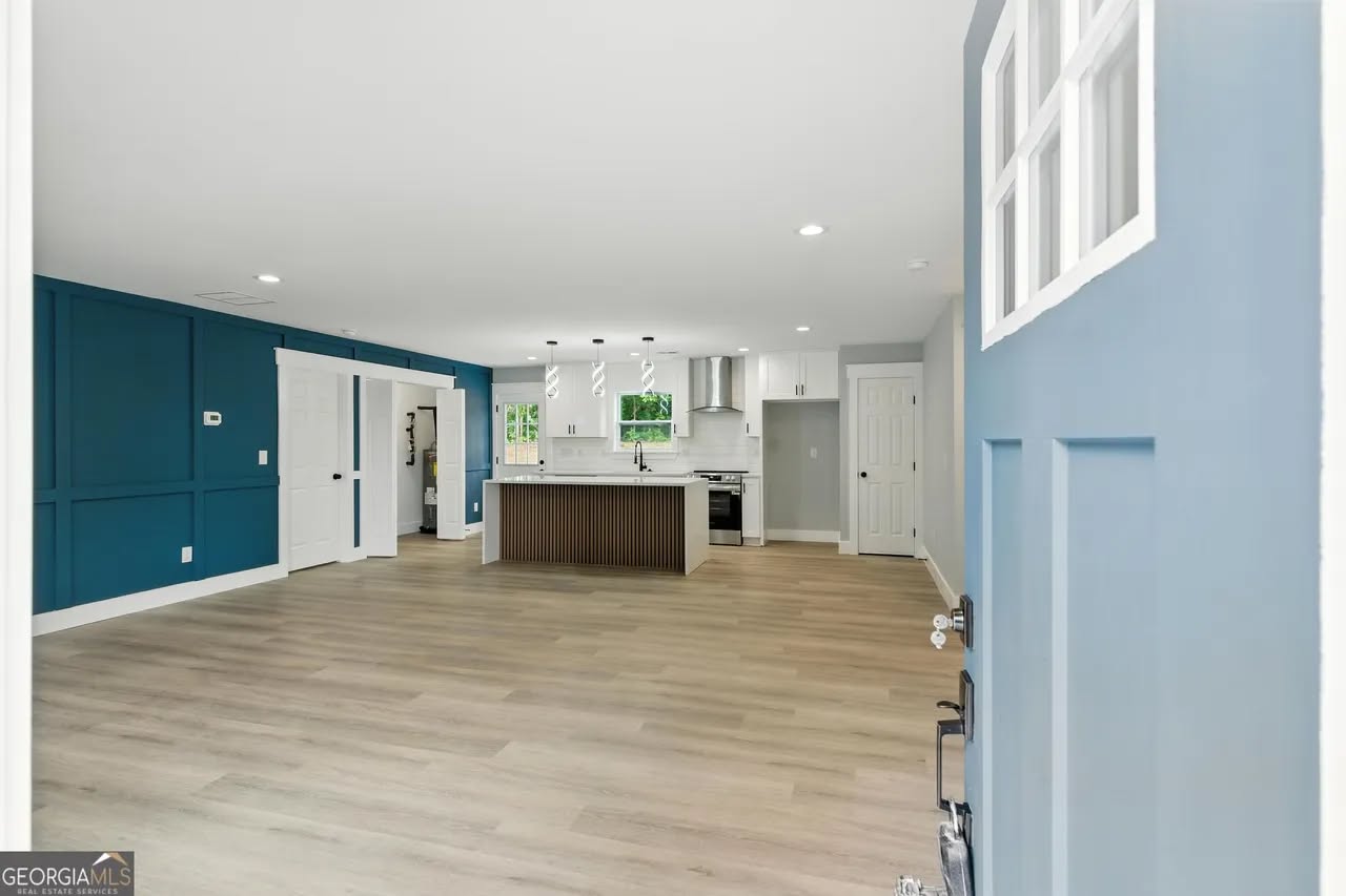 Spacious modern living area with light wood floors, blue accent wall, and an open kitchen featuring an island and stainless steel appliances.