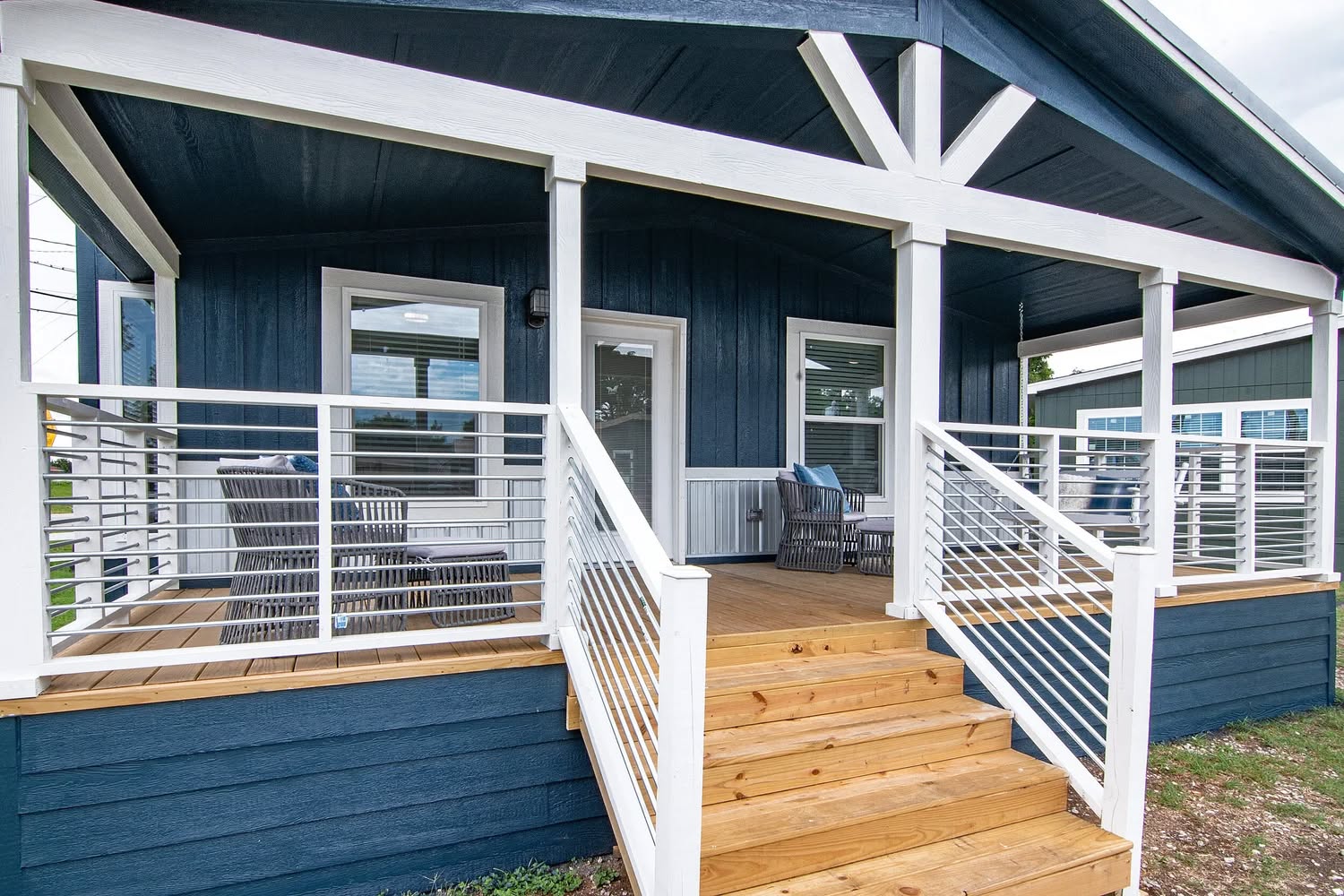 Front porch of a blue house with white trim, featuring wooden steps and railing. Two sets of wicker chairs create a welcoming, cozy atmosphere.