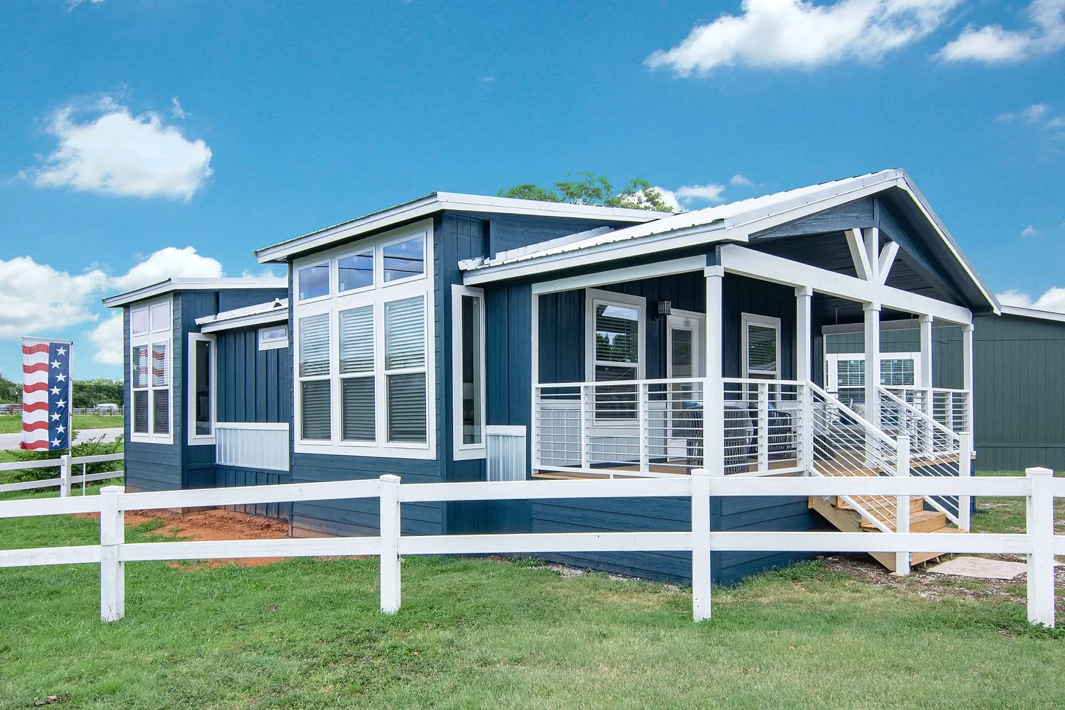 A modern blue manufactured home with white trims and large windows, surrounded by a white fence. The scene is bright and clear under a blue sky.