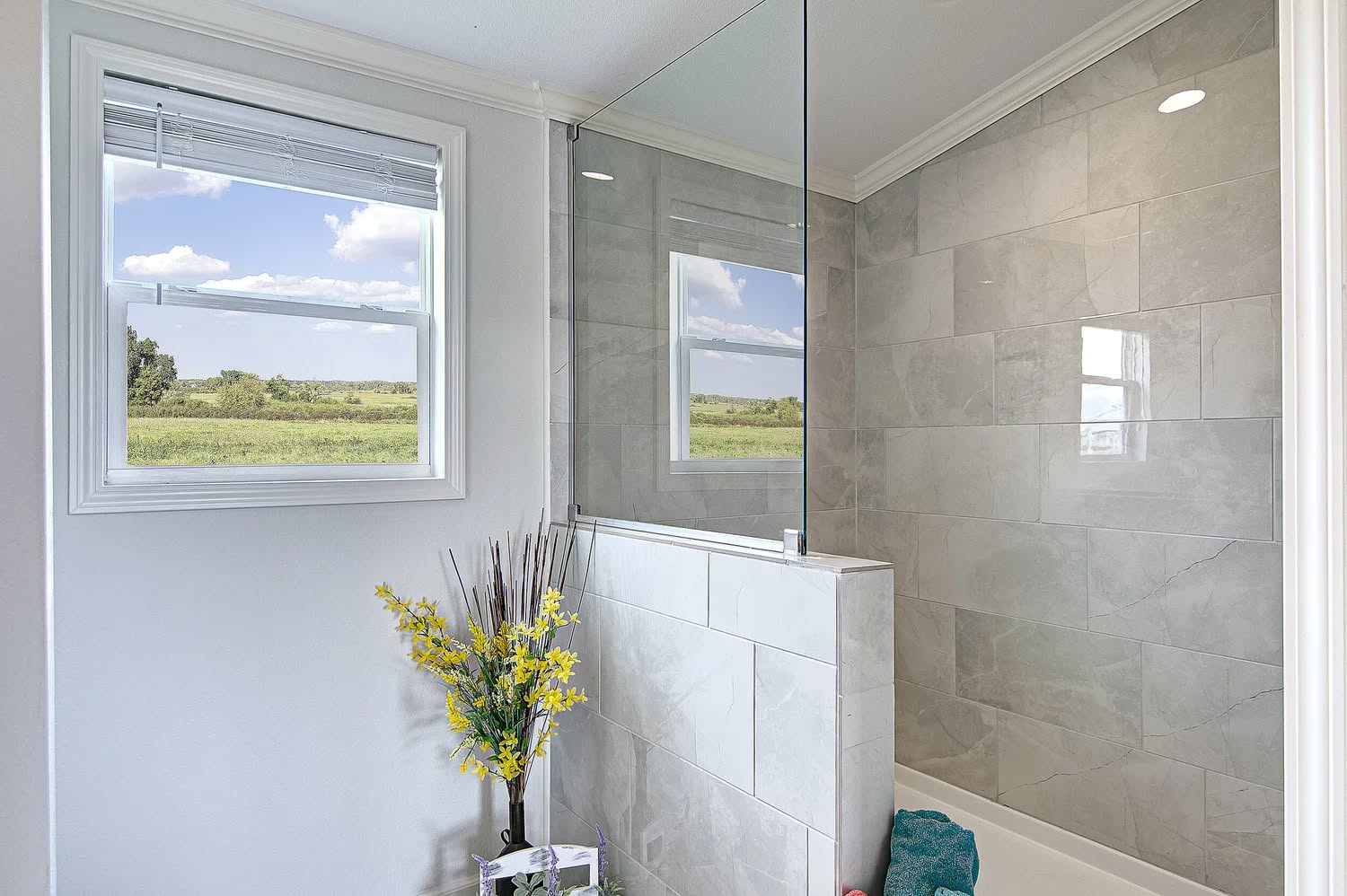 Bright bathroom with large tiles, a glass shower, and a window showcasing lush greenery. Yellow flowers in a vase add a touch of nature.