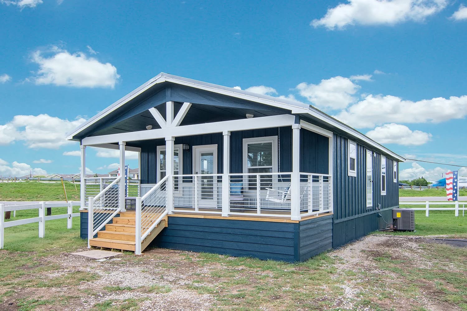 Small blue house with a white railing porch sits under a clear sky. Wooden steps lead up to the entrance, surrounded by green grass and open landscape.