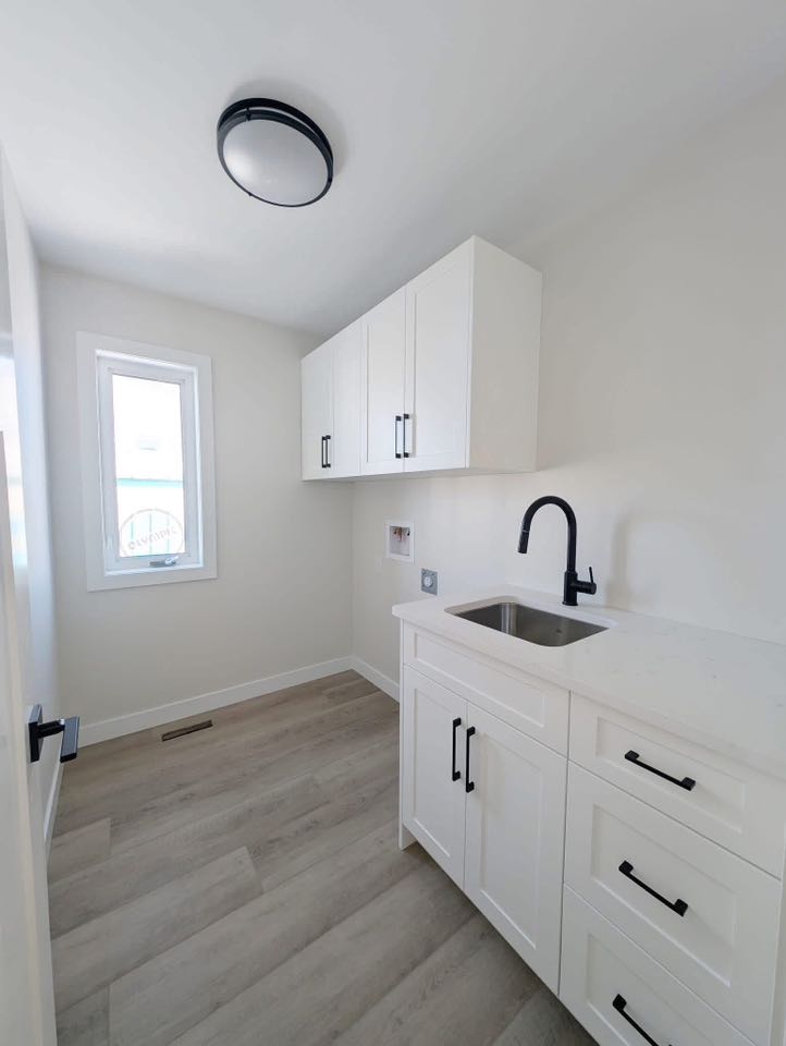 Minimalist laundry room with white cabinets and a sleek black faucet. Natural light from a window highlights the light wood flooring. Clean and modern ambiance.
