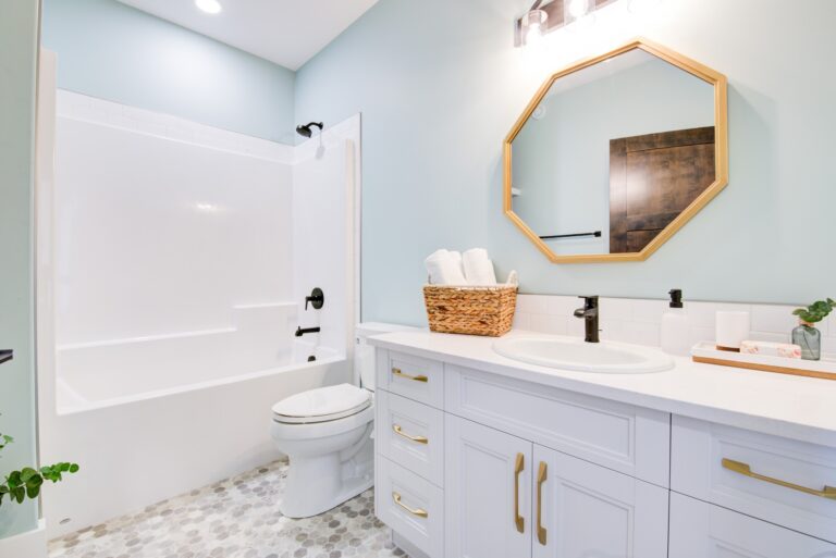 A modern bathroom with light blue walls, hexagonal mirror, white vanity with gold handles, basket with towels, and white bathtub. Bright and clean ambiance.