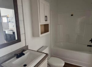 Modern bathroom with white walls, a bathtub, sink with dark faucet, toilet, and wooden mirror. Cabinets have open shelving. Calm, minimalist vibe.