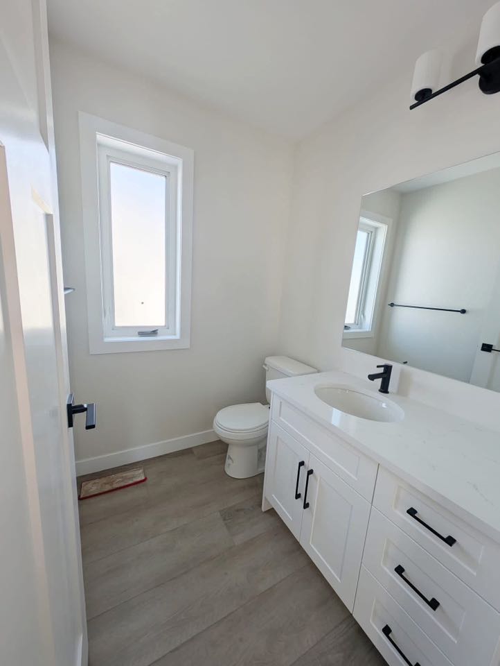 Bright, minimalist bathroom with white walls and cabinets, a black faucet, oval sink, and large mirror. Natural light from a tall window.