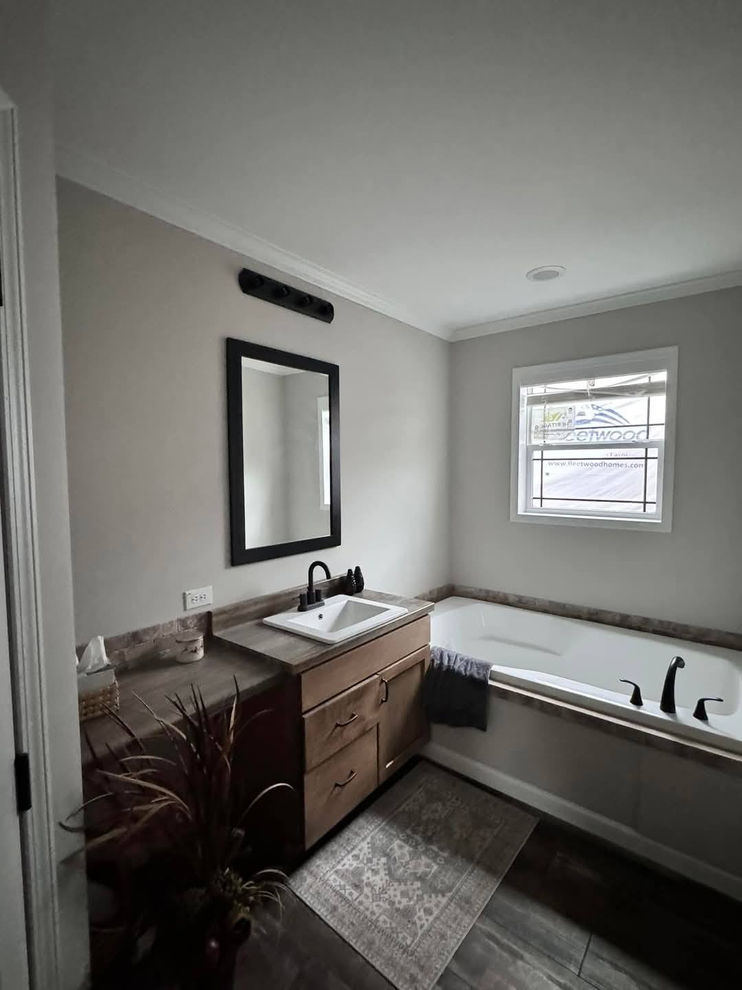 A modern bathroom with gray walls features a large window, white bathtub, wooden vanity with a sink, and a black-framed mirror, creating a calm atmosphere.