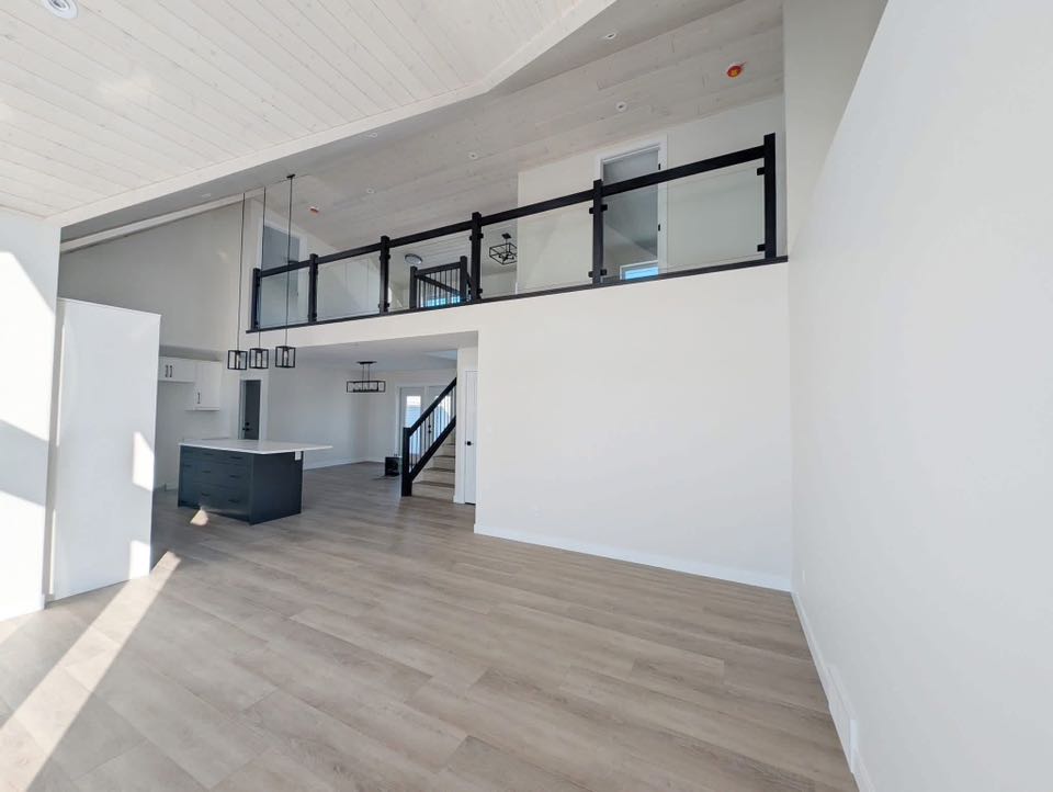 Spacious modern interior with high ceilings, light wood flooring, and white walls. An upstairs balcony with glass railing overlooks an open kitchen.