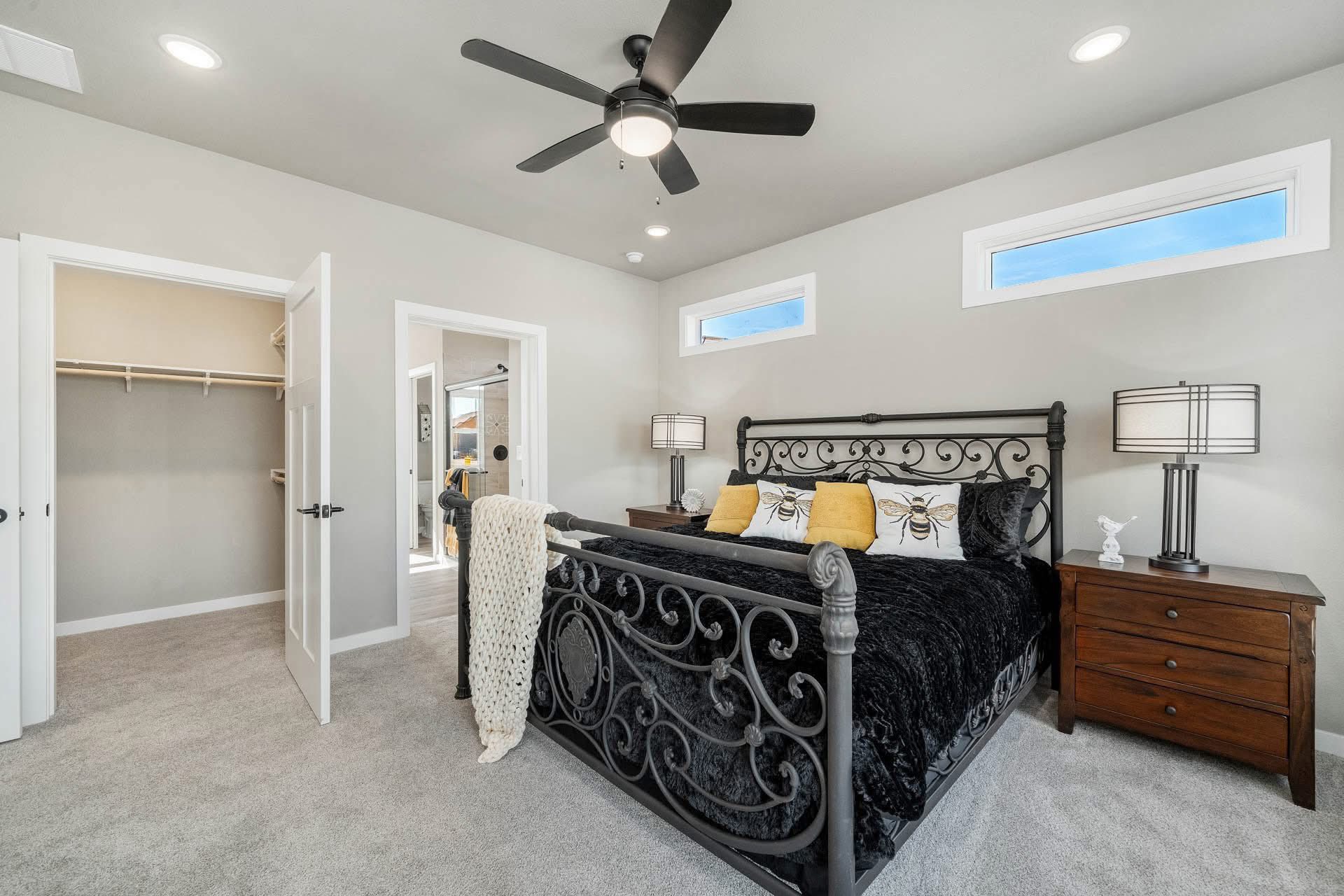 Bedroom with an ornate black metal bed, black and yellow pillows, cozy gray carpet, wooden nightstands, and ceiling fan. Open closet and bathroom doors add depth.