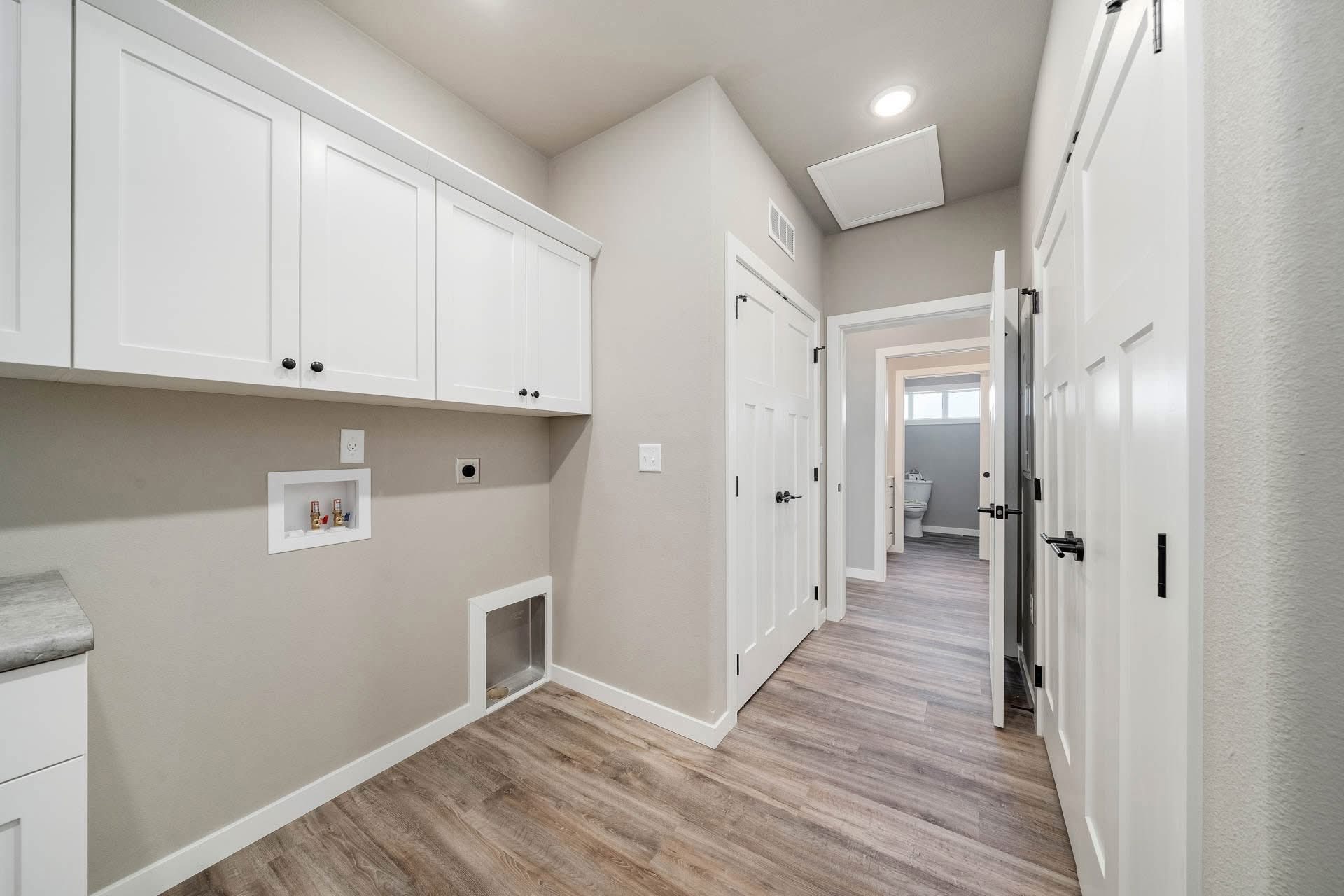 Bright laundry room with white cabinets, wood flooring, and beige walls. Open doorway leads to another room, creating a spacious and clean atmosphere.