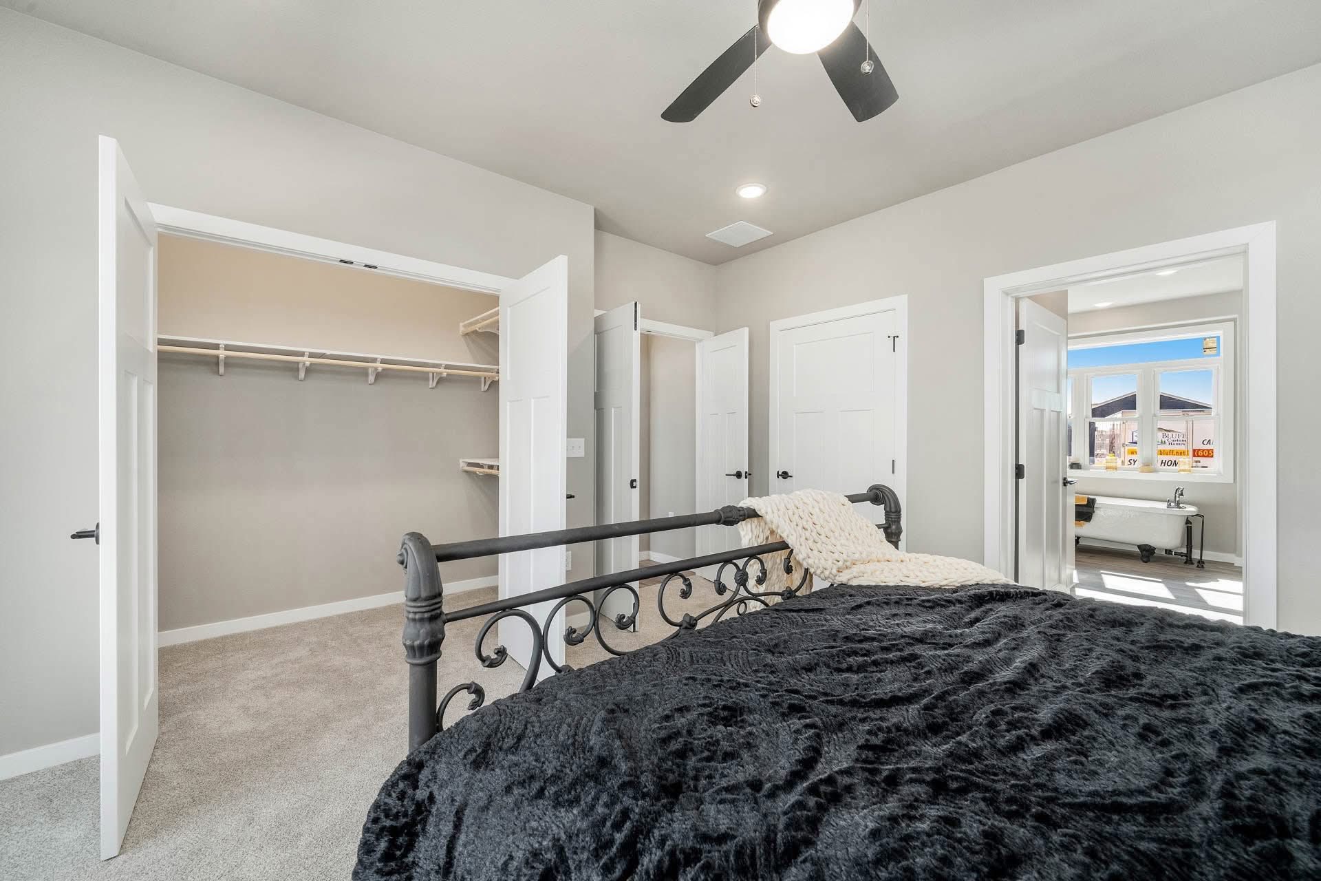 Bedroom with a black metal bed, plush dark comforter, and soft lighting. Open doors reveal a spacious closet and bathroom. Calm and inviting atmosphere.