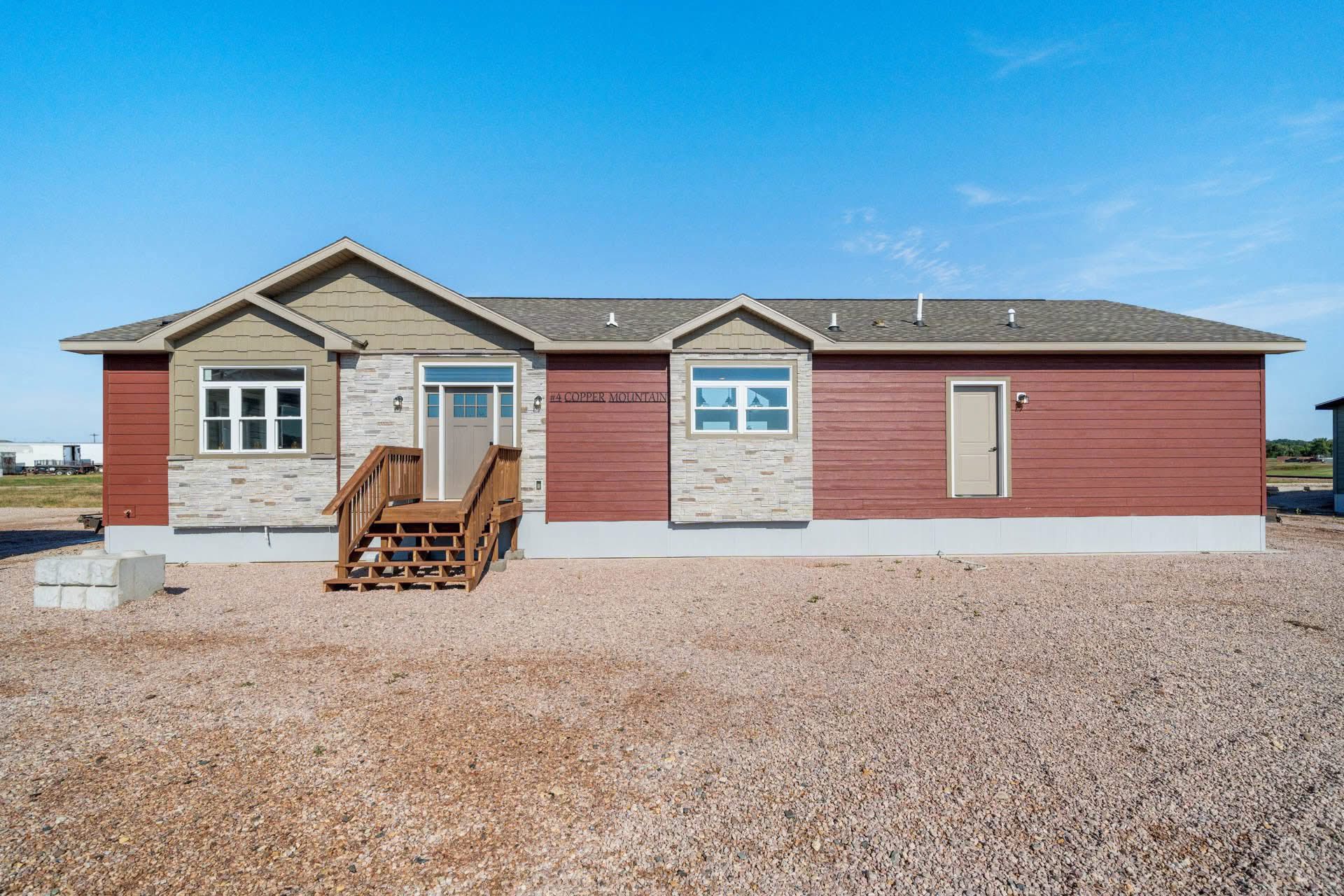 Single-story red and beige house with stone accents, wooden steps, and a gable roof. It's set against a clear blue sky and gravel foreground.