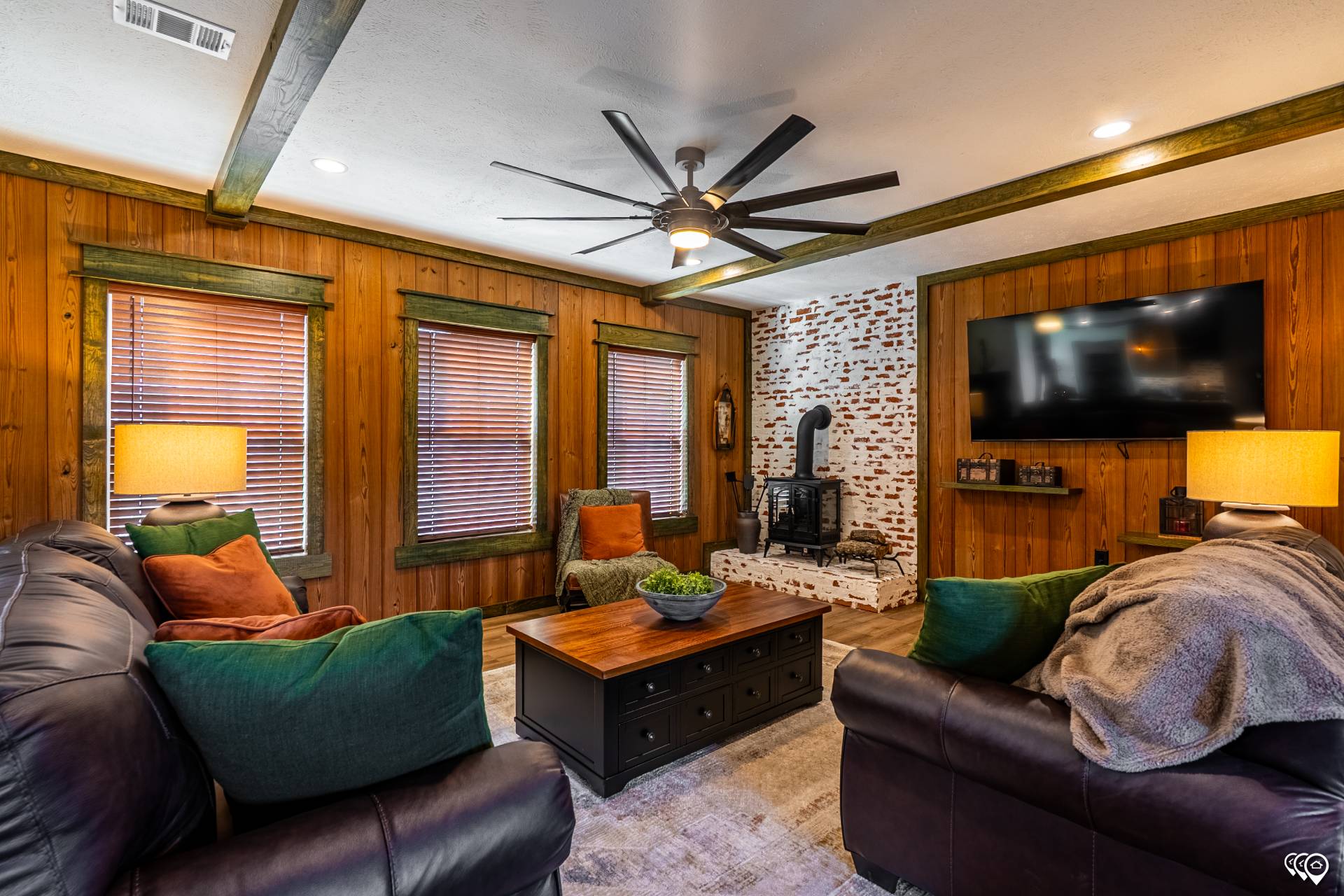 Cozy living room with wooden walls, leather sofas, colorful cushions, and a ceiling fan. A rustic stove is set against a white brick wall under a mounted TV. Warm lighting from lamps creates a welcoming atmosphere.