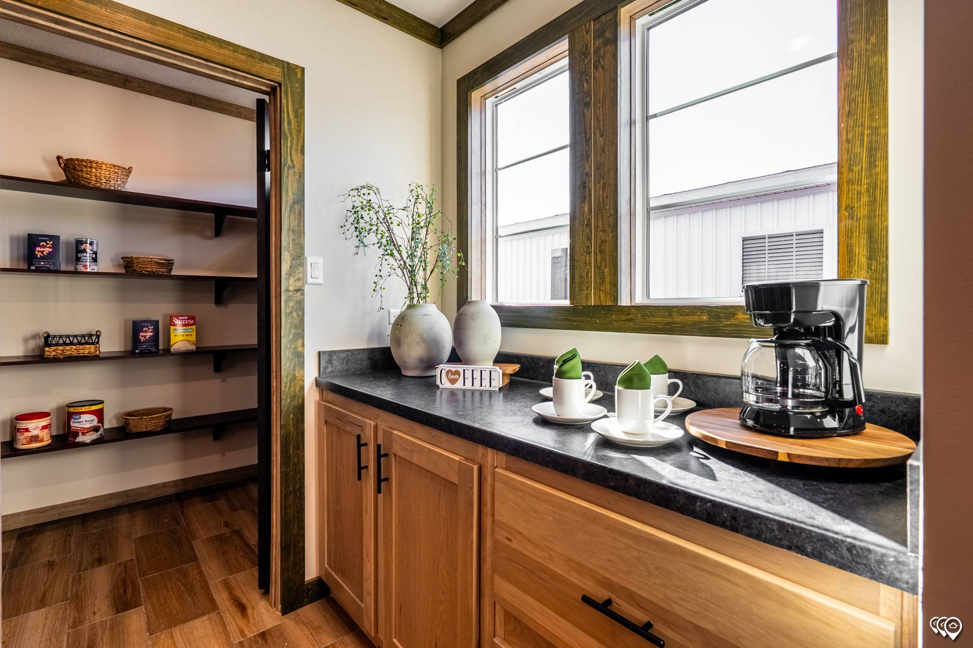 A cozy kitchen corner with natural wood cabinets and a black countertop. A coffee maker, cups, and green napkins are set. A pantry with items is visible.
