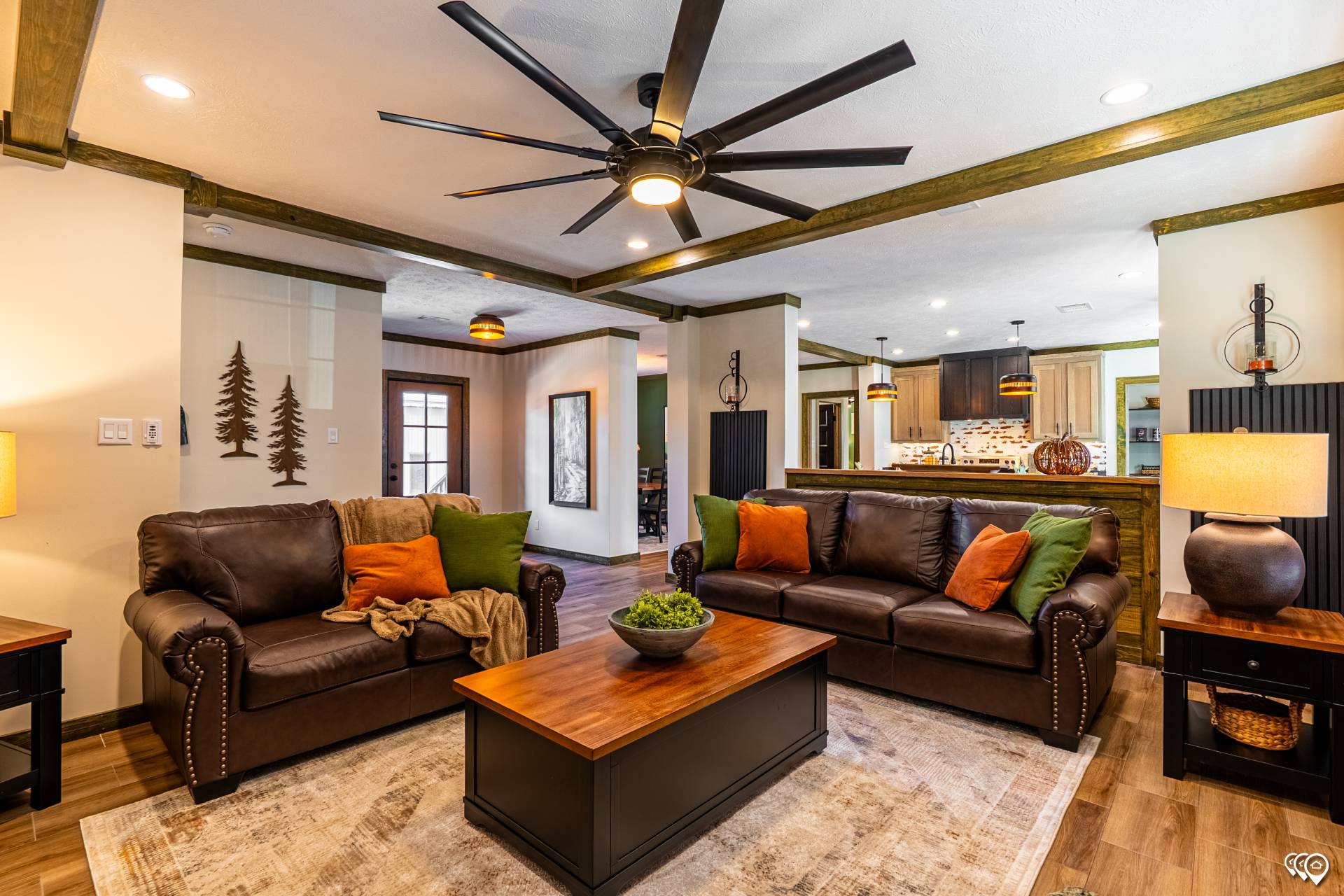 Cozy living room with two brown leather sofas adorned with orange and green pillows. A wooden coffee table holds a plant centerpiece. Warm lighting, large ceiling fan, and tree decorations create a rustic, inviting atmosphere.
