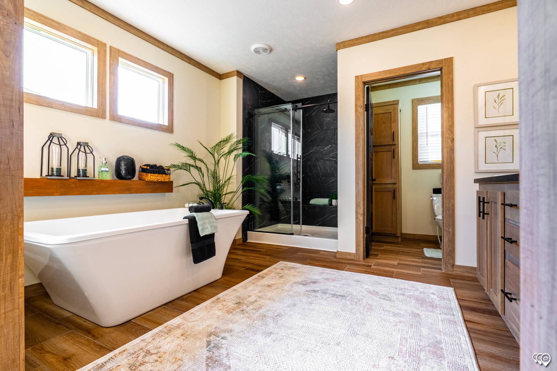 Spacious bathroom with a sleek white bathtub, black-tiled shower, wooden accents, and large beige rug. Natural light and decor create a serene ambiance.