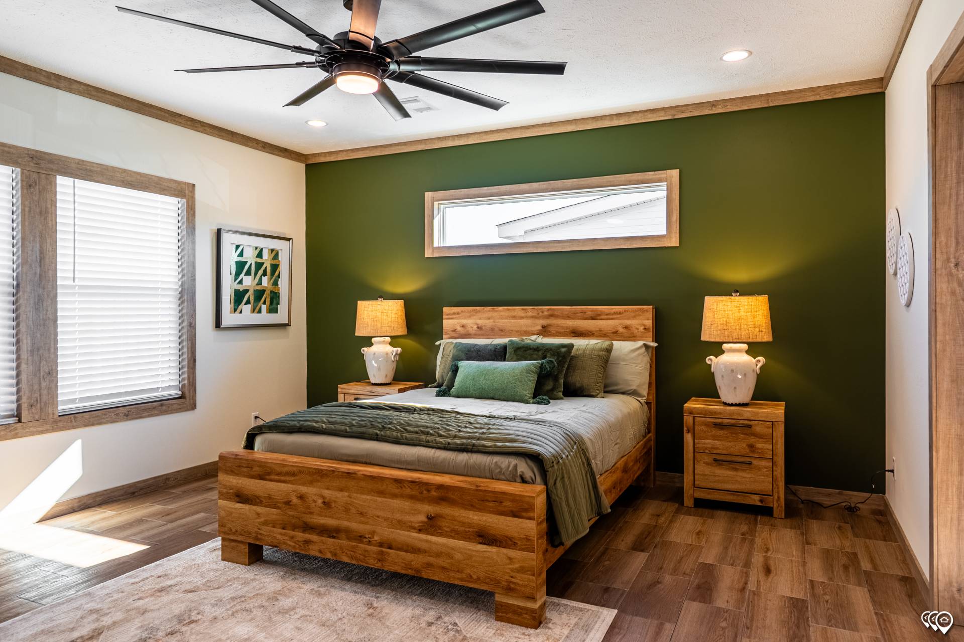 A cozy bedroom features a wooden bed with green and gray bedding against an olive accent wall. Matching lamps on nightstands and large ceiling fan add warmth.