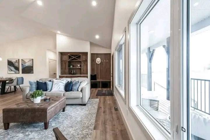 Spacious living room with a cozy beige sofa, blue and white cushions, a brown ottoman, large windows, a wooden floor, and a built-in bookcase.
