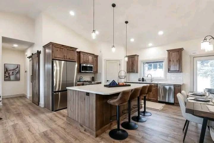 A modern kitchen features a wooden island with three brown stools, stainless steel appliances, and wooden cabinets. The space is bright and welcoming.