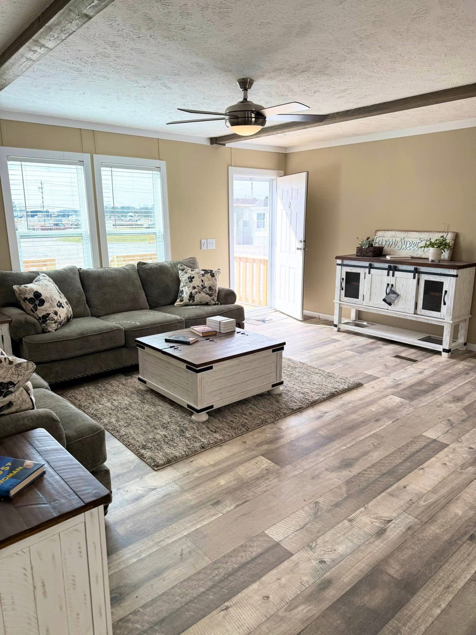 Bright living room with beige walls, light wood flooring, and a gray sofa adorned with floral cushions. A rustic coffee table on a rug sits centrally. An open door lets in natural light, enhancing a calm, inviting mood.