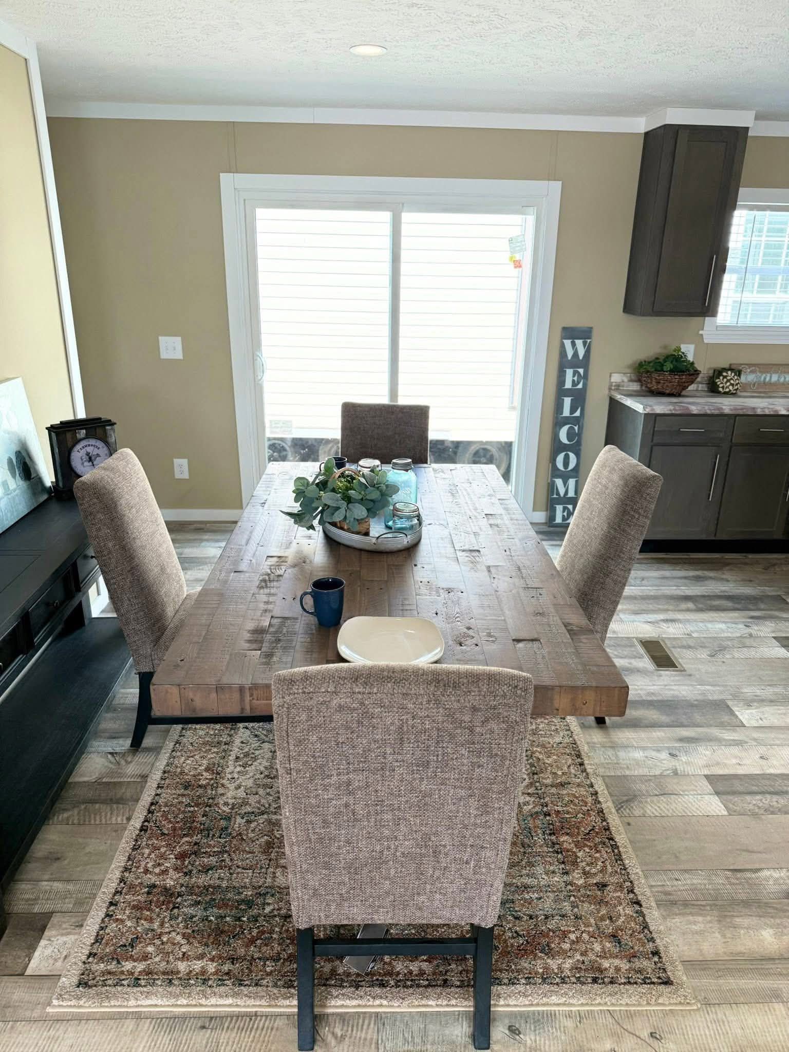 A dining room with a wooden table, four upholstered chairs, and a patterned rug. A welcome sign stands near sliding glass doors, conveying a warm and inviting atmosphere.