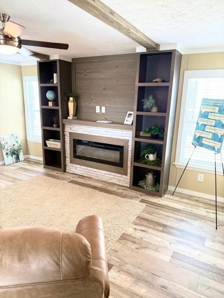 Cozy living room with a modern electric fireplace set in a white brick mantle. Brown bookshelves flank the fireplace, holding decor items, plants, and a globe. A ceiling fan hangs above, and light wood flooring underscores the warm ambiance.