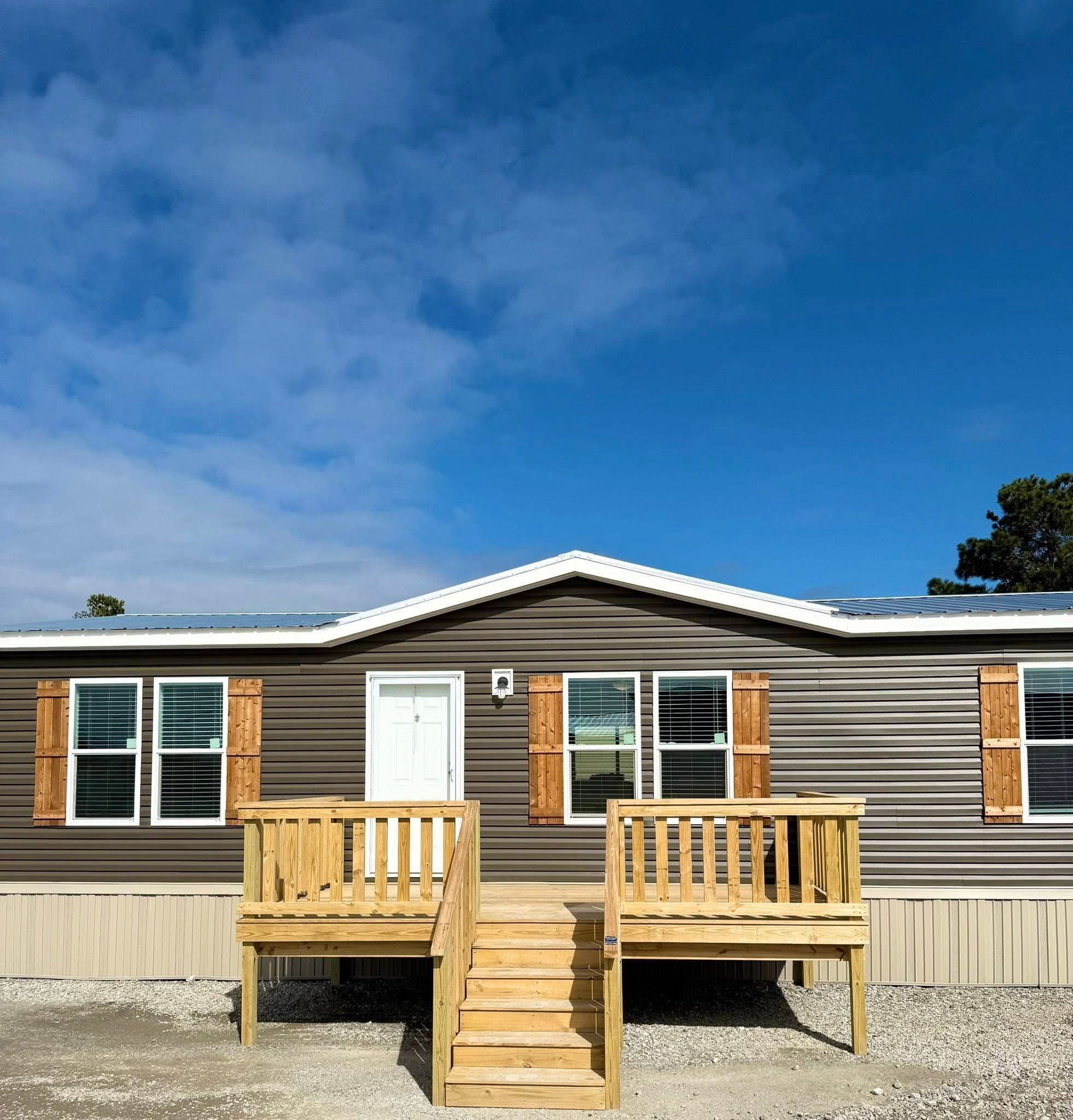 A single-story brown mobile home with white trim and wooden shutters under a bright blue sky. A small porch with stairs leads to the front door.
