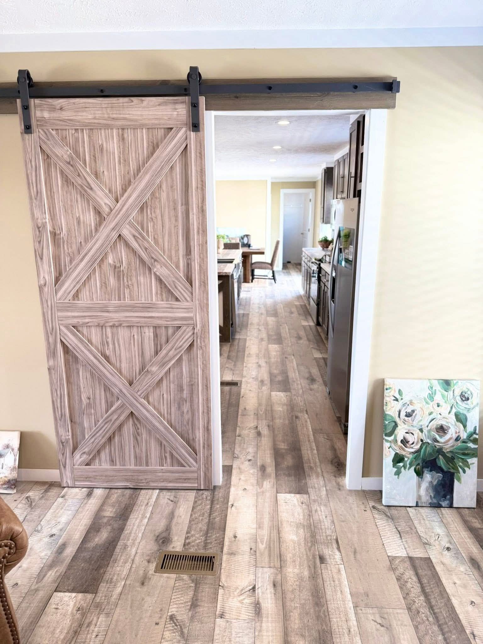 A rustic sliding barn door partially open, revealing a hallway with wooden floors leading to a bright kitchen. A floral painting leans against a cream wall.
