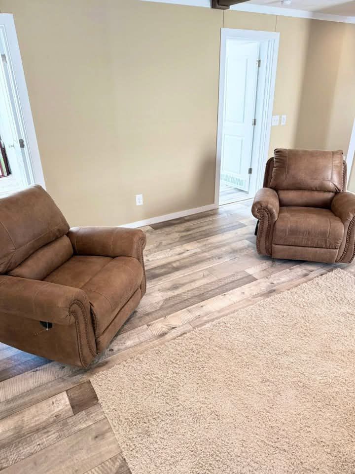 Two brown recliners sit on a hardwood floor near a beige wall and a textured carpet. The room is sparsely decorated, conveying a cozy, minimalist vibe.
