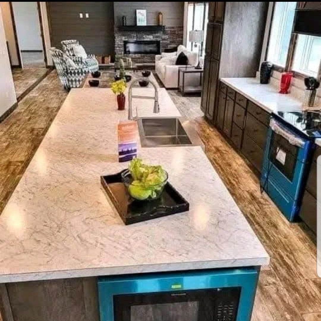 Modern kitchen with a long marble island, sink, decorative plants, and a bright blue oven. Cozy living room in the background exudes warmth.