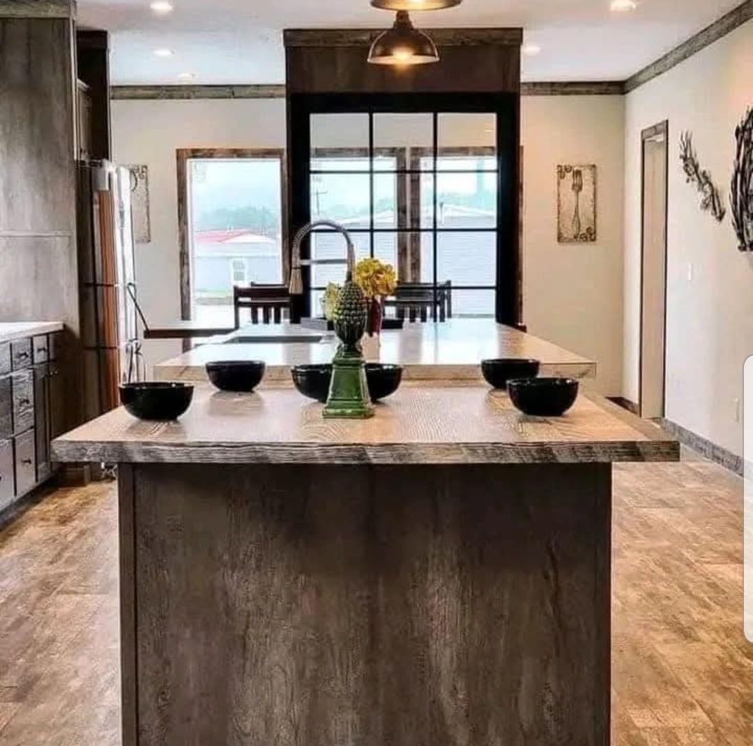 A rustic kitchen features a large wooden island with four black bowls and a green vase with flowers. Earth-toned decor creates a cozy, inviting feel.