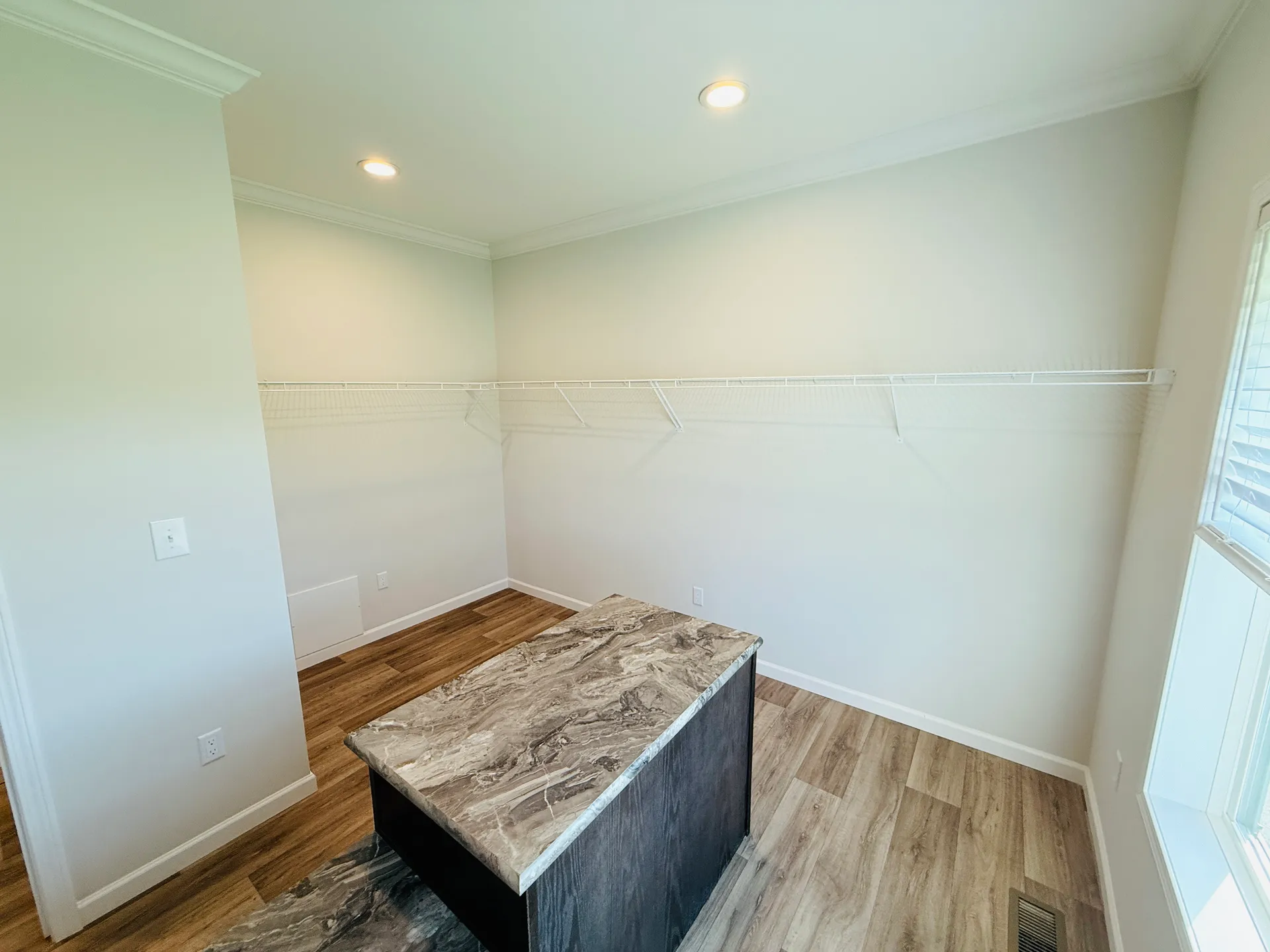 A minimalist walk-in closet with light beige walls, wood flooring, and an island with a marbled top. Wire racks line the walls for storage.