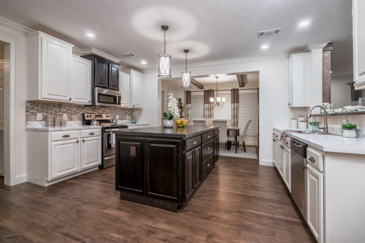 Spacious kitchen with dark island, white cabinets, and stainless steel appliances. Bright lights, wooden floor, and dining area in the background.