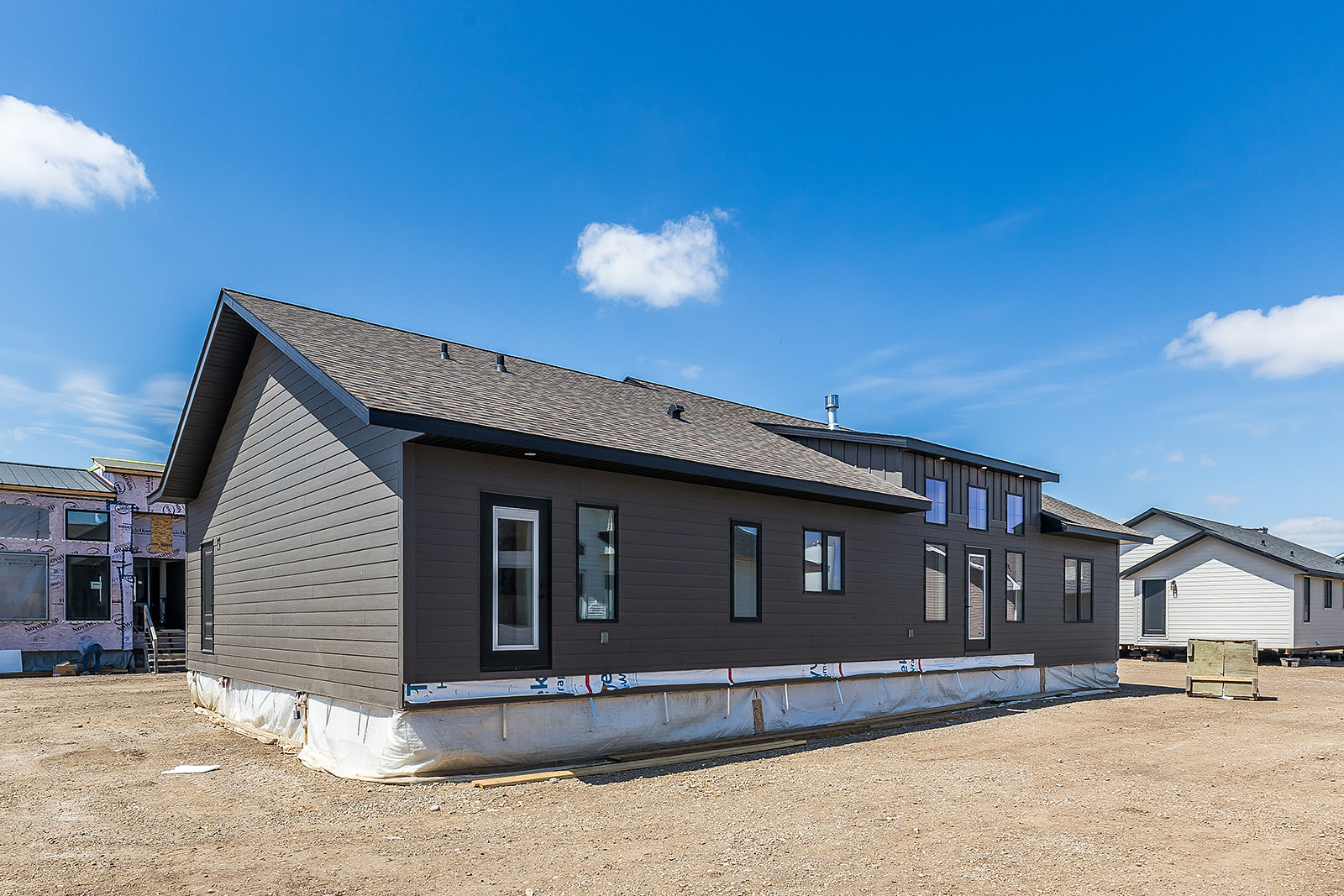 Newly constructed gray modular home on a construction site under a clear blue sky. The building has several windows and a shingled roof.