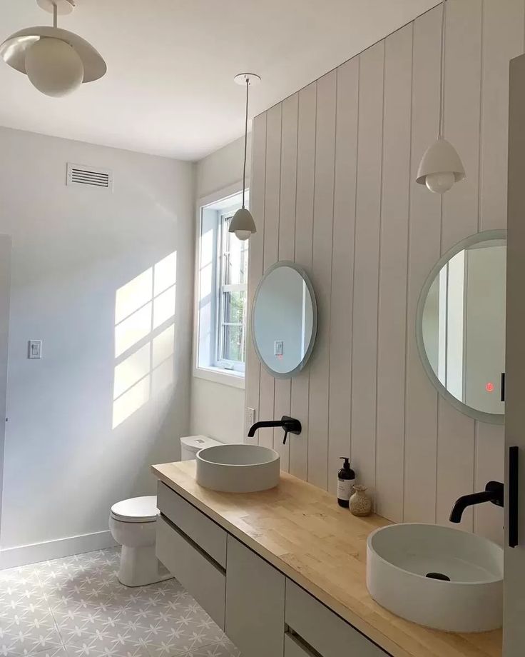 Modern bathroom with light wood countertops, dual round mirrors above vessel sinks, black faucets, and pendant lighting. Soft natural light enters through a window, creating a calm atmosphere.