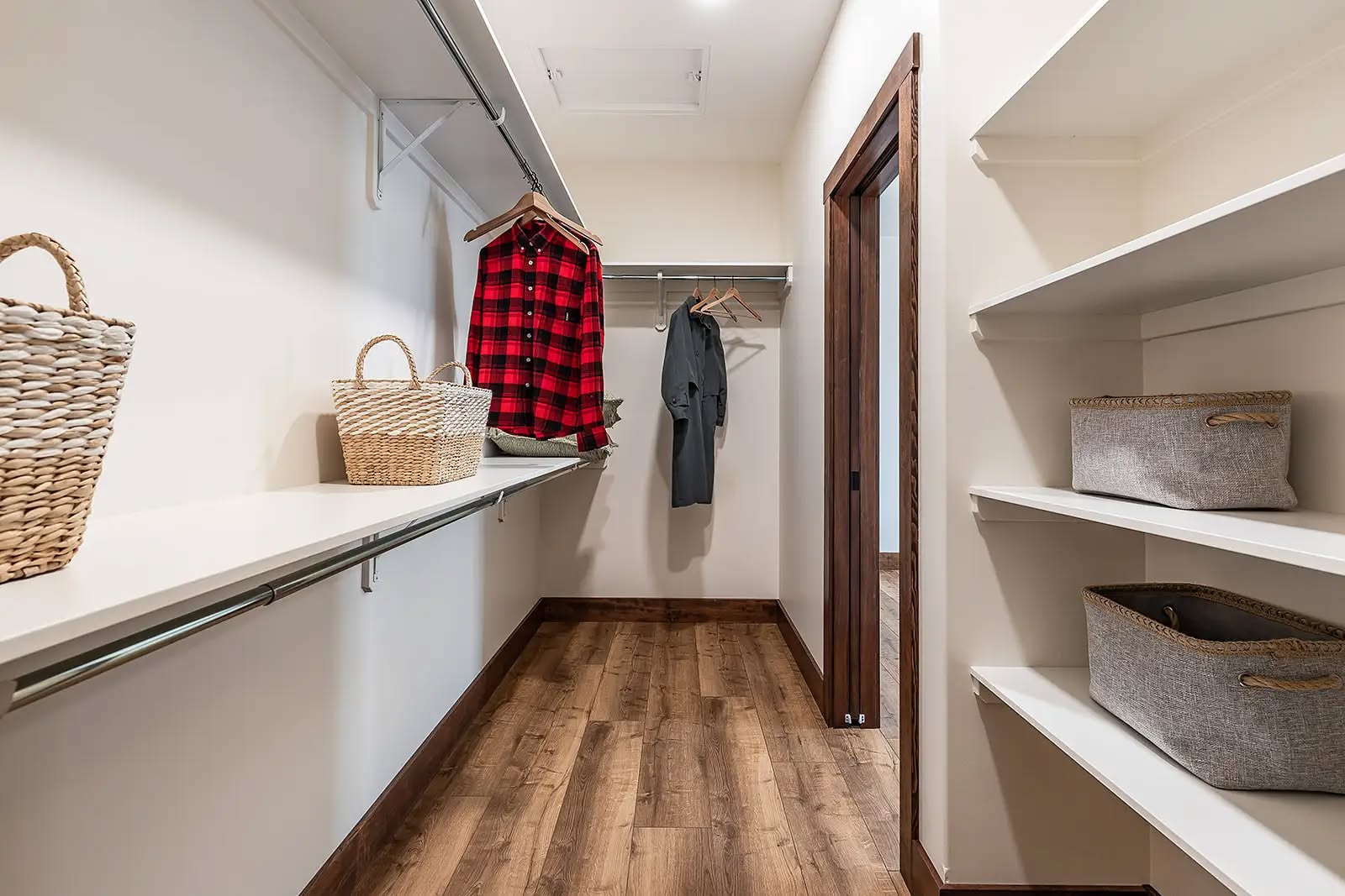 Walk-in closet with wooden flooring, featuring hanging red plaid and gray shirts, wicker baskets on shelves, and a minimalist, organized design.