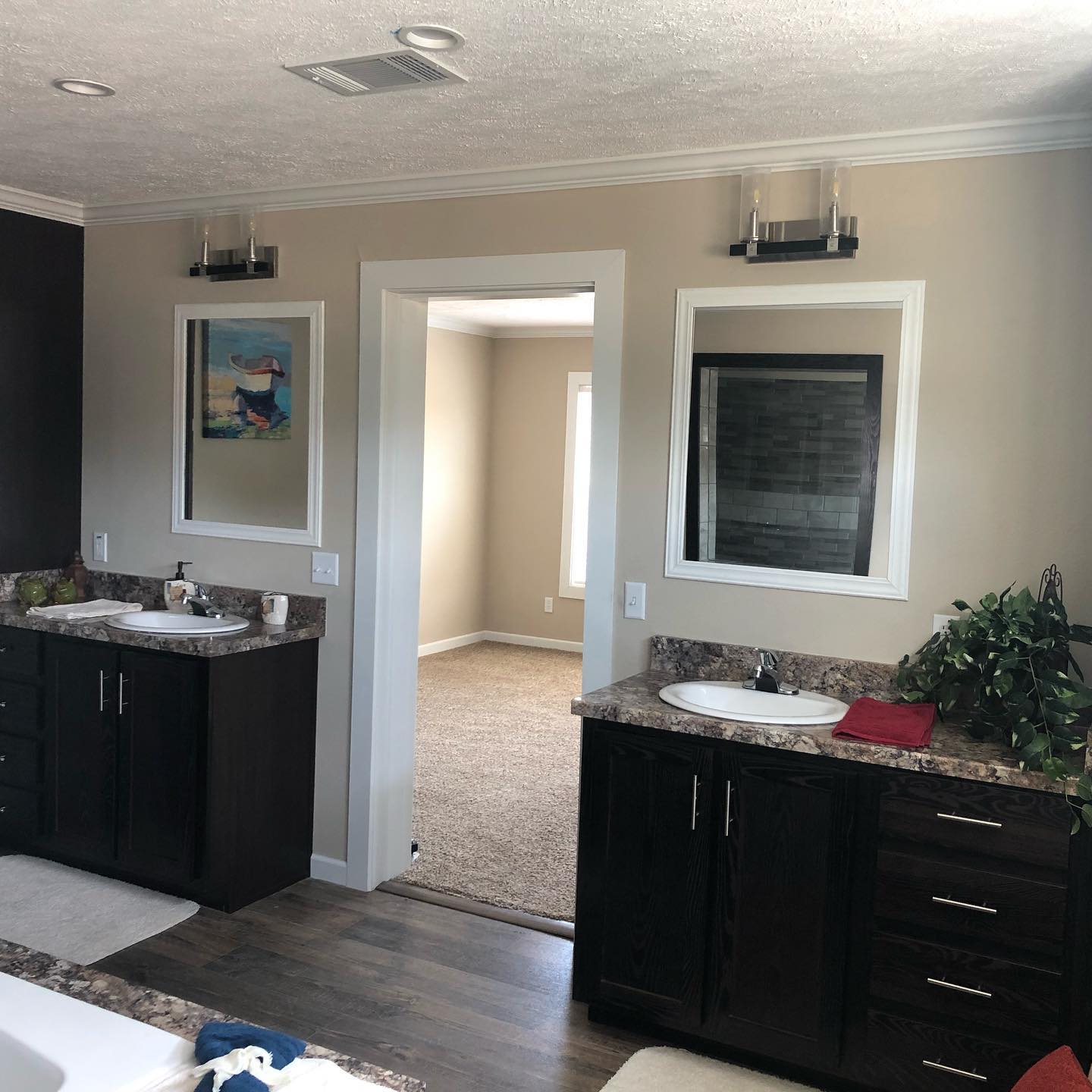 Modern bathroom with dark wood double vanities, granite countertops, and framed mirrors. Soft lighting, wall art, and a doorway to a carpeted room.