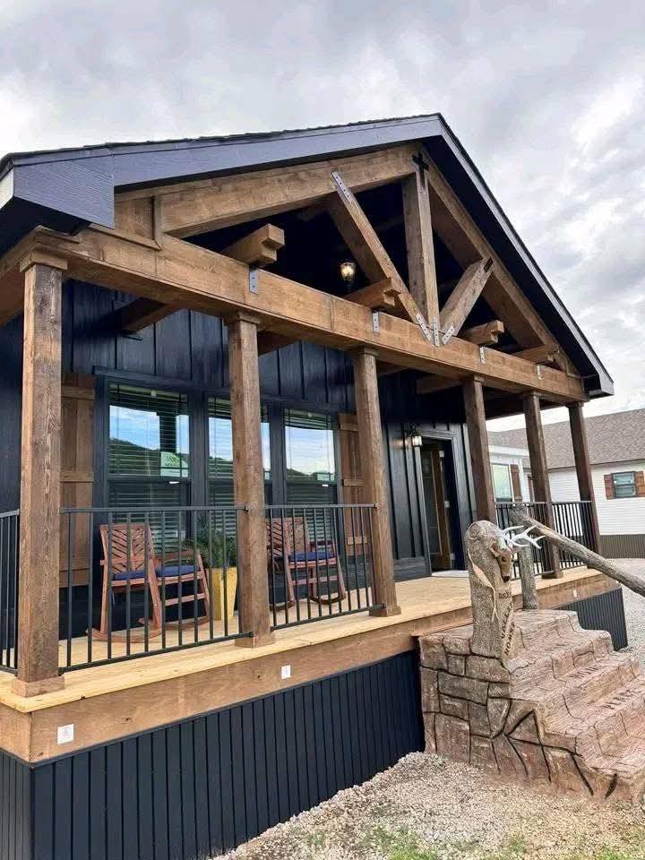 A rustic cabin with a wooden porch featuring exposed beams and pillars. Two chairs sit on the porch, and a stone water pump is in the foreground. Cloudy sky.